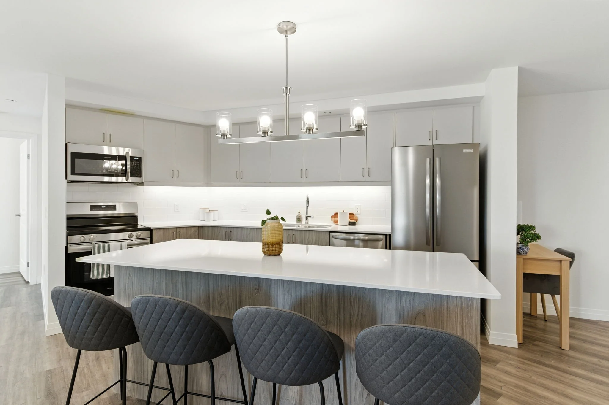 Modern kitchen in St. Thomas, ON rental apartment with white and gray cabinetry, stainless steel appliances, a large white island with four gray quilted chairs, a chandelier, and a small wooden table with a plant in the background.