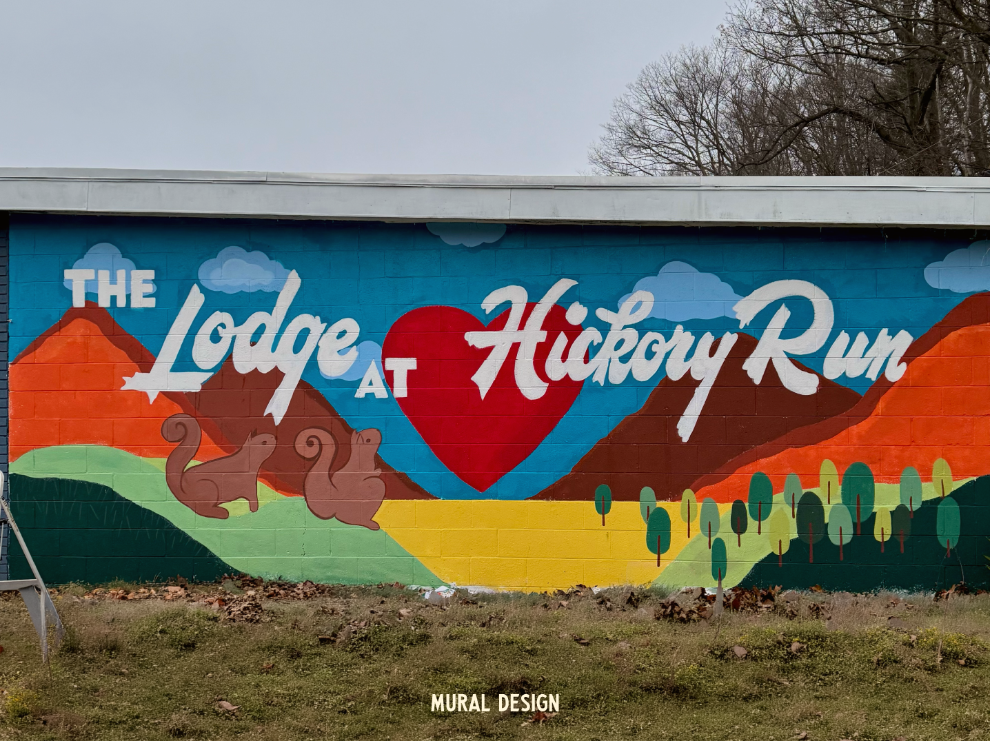 A colorful mural on a brick wall advertising 'The Lodge at Huckleberry Run' with mountains, trees, clouds, a large red heart, two hummingbirds, and leaves painted in a landscape style.
