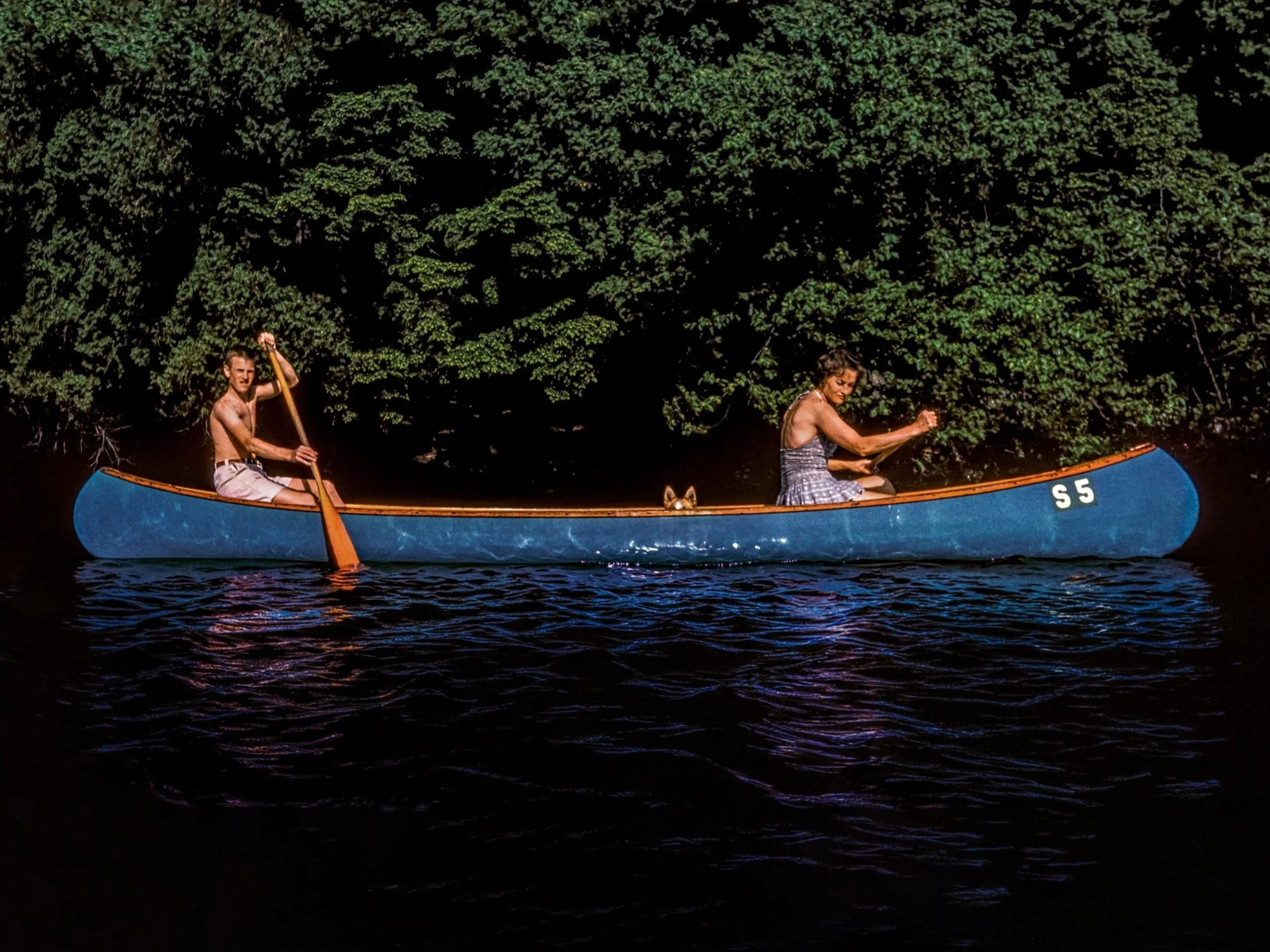 A young man and a woman paddle a blue canoe on a dark lake, with a background of dense green trees.