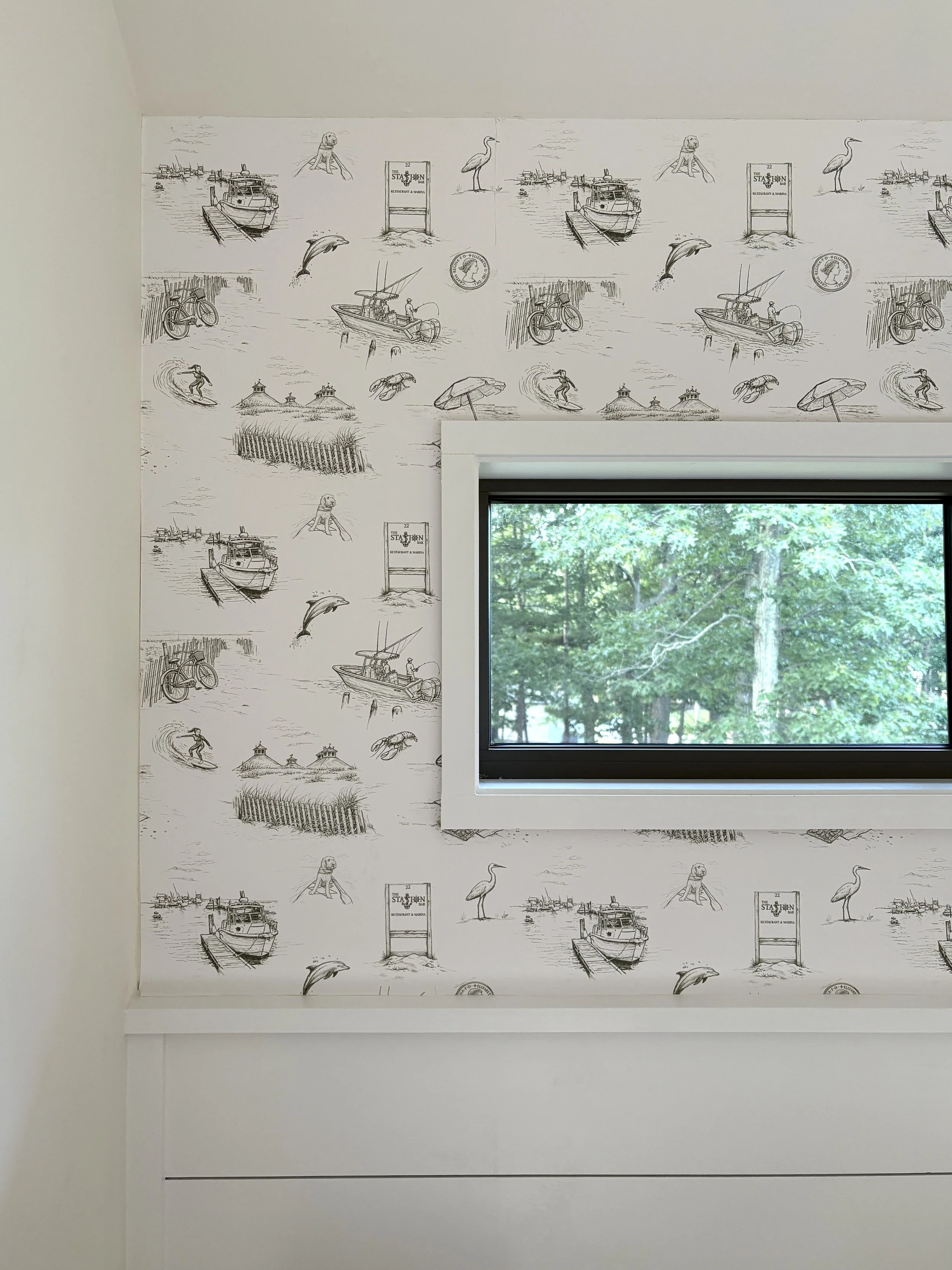 The image shows a room with a wallpaper featuring black and white sketches of boats, a bicycle, a dolphin jumping out of the water, a person surfing, a lighthouse, and various other seaside scenes. There is a rectangular window showing green trees outside.
