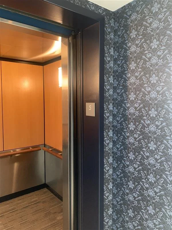 Open elevator door revealing wood panel interior with metal accents, next to a wall with blue floral wallpaper, and a small sign with the number 5.