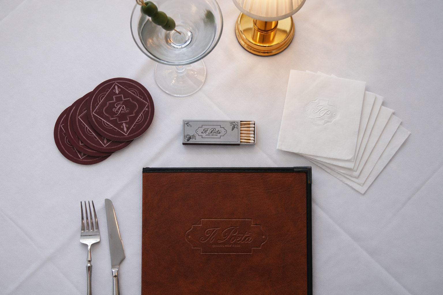 Table setting with a white tablecloth, a brown menu, silverware, napkins, a glass of water with green olives, a box of matches, and a brass lamp.
