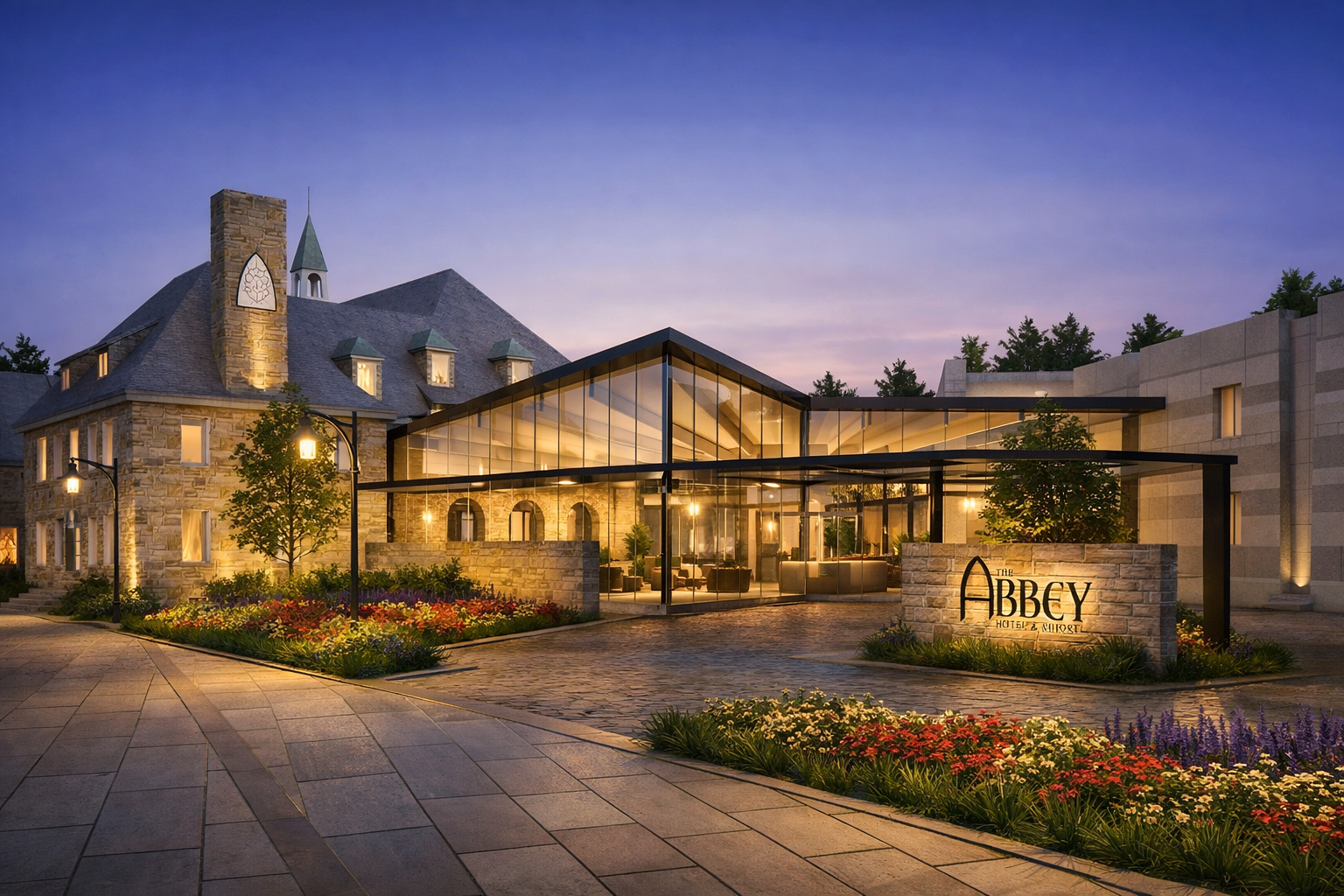 Exterior view of The Abbey Hotel & Resort during dusk, showcasing a modern glass entrance with warm lighting, flowerbeds, and a historic stone building in the background.