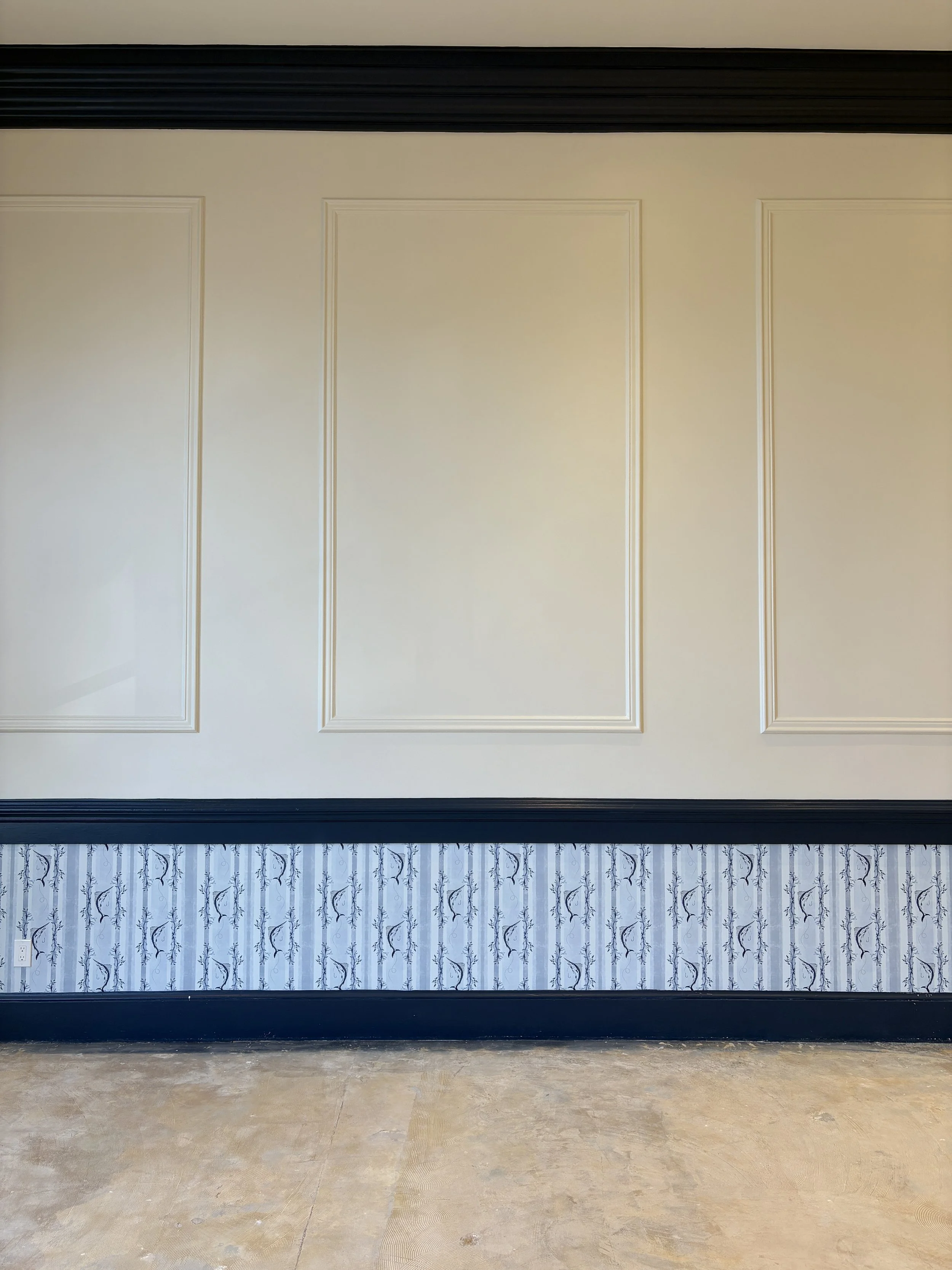 Interior wall painted in cream with decorative molding and patterned wallpaper below, separated by a black chair rail, and a concrete floor.