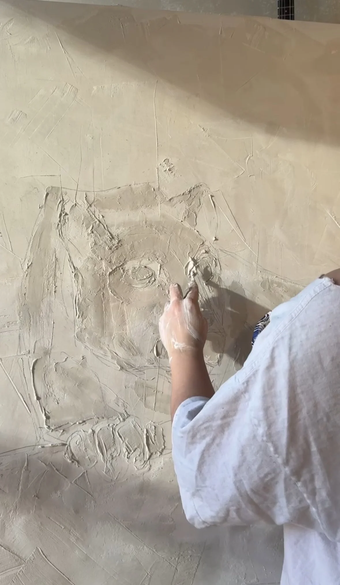 Person carving a face into a beige plaster wall with a small tool, wearing a white shirt.