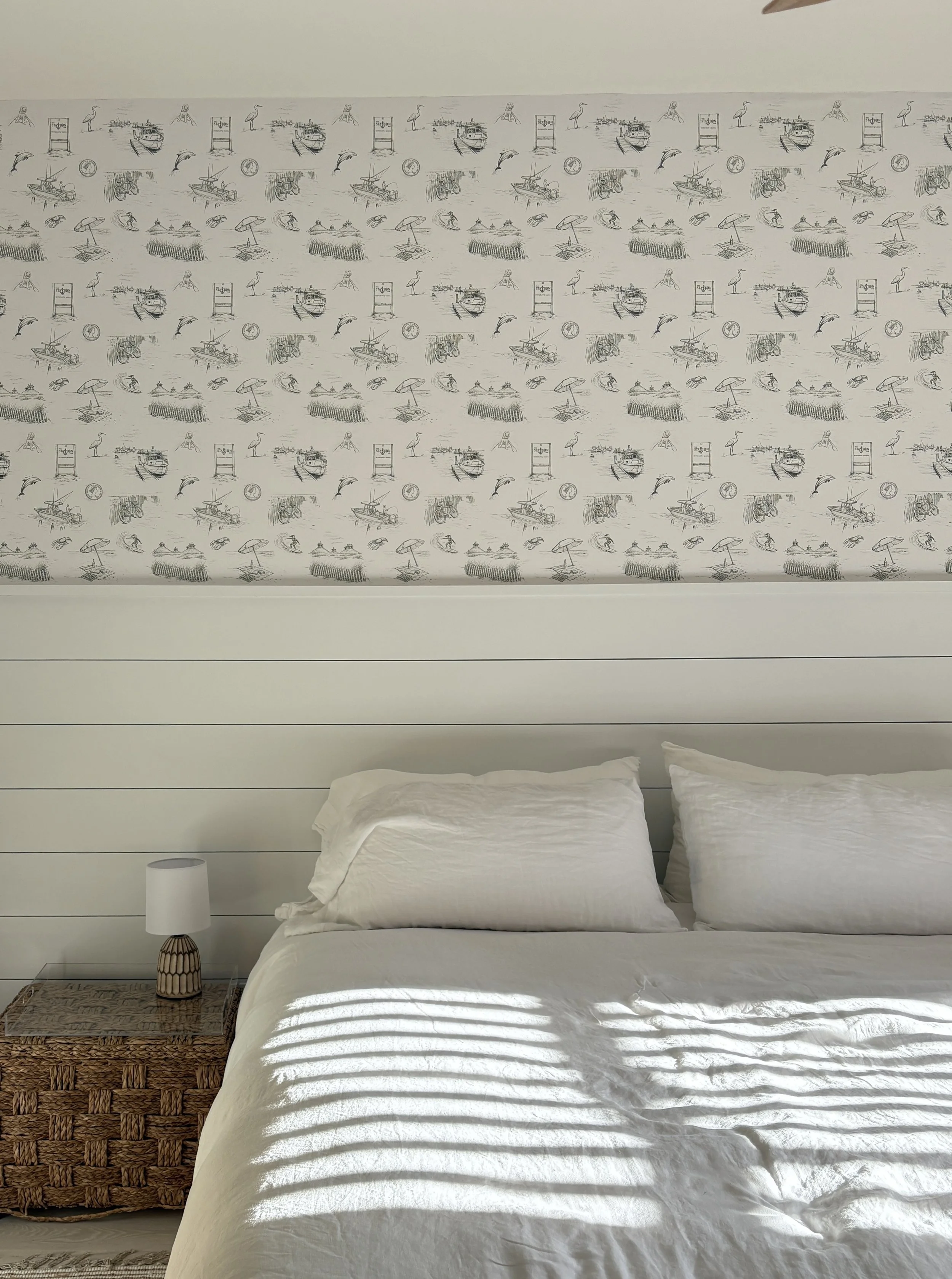A bedroom with a white bed, a rattan nightstand, a white lamp, and a decorative wall panel with black and white sketches of boats, a heron, dolphins, and tropical scenes.