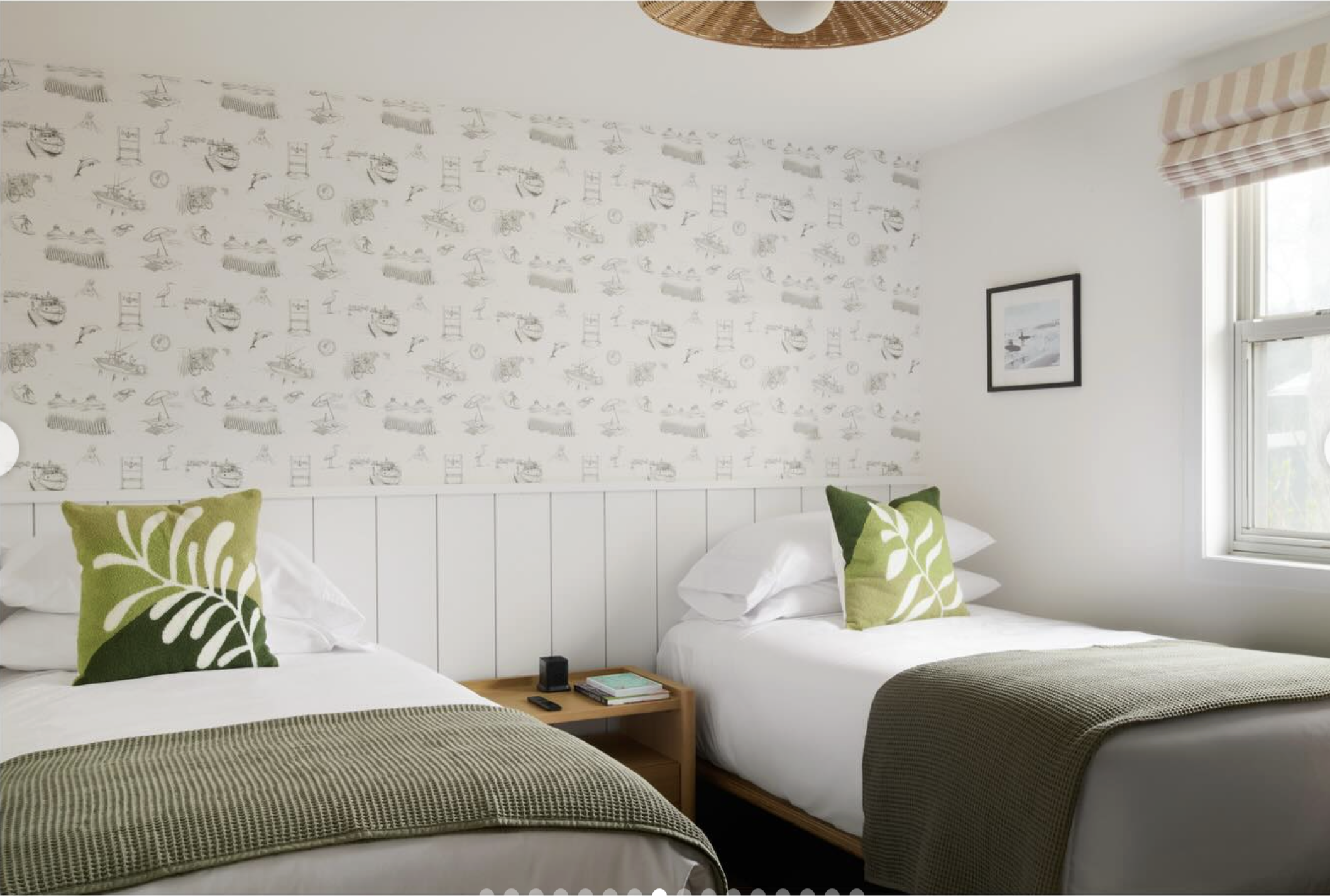 A bedroom with two beds, each with green and white pillows, a wooden nightstand with a book and remote, a framed black-and-white photograph on the wall, and window with striped valance curtains