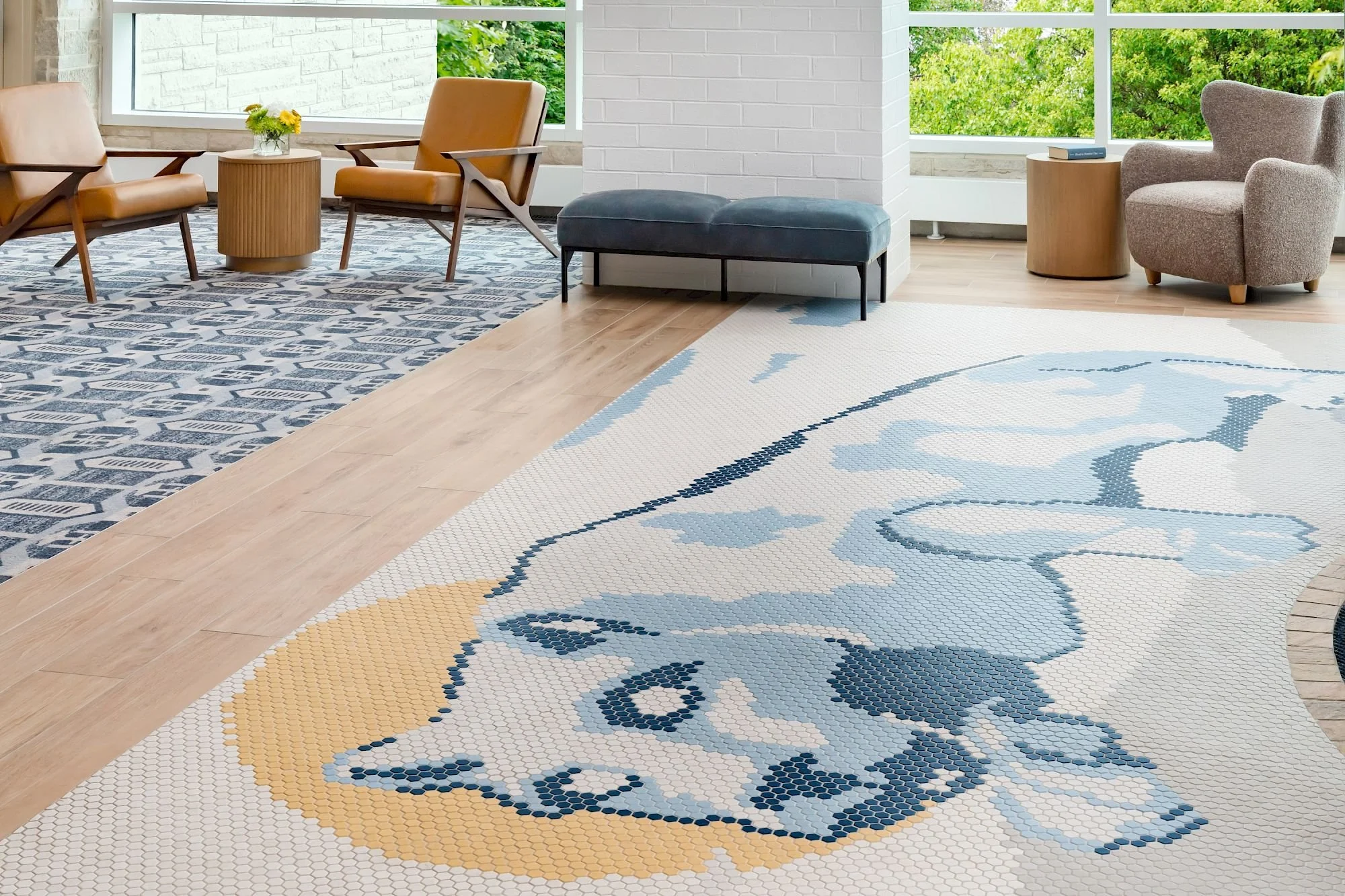 A room with large windows, featuring a seating area with two brown leather chairs, a gray armchair, a blue bench, and a patterned blue and white rug with a unicorn design and a yellow detail.