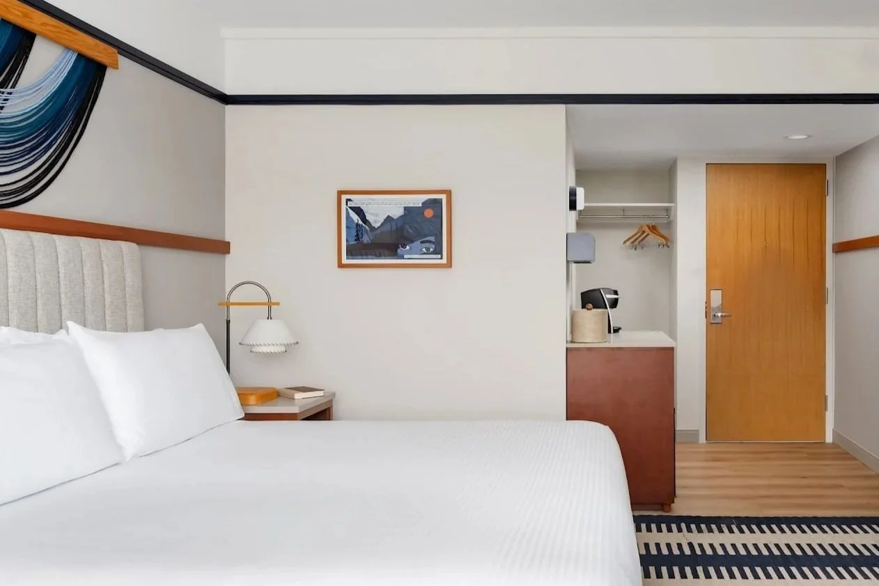 A hotel room with a bed, bedside table, wall-mounted lamp, framed artwork, kitchenette, and entrance door.