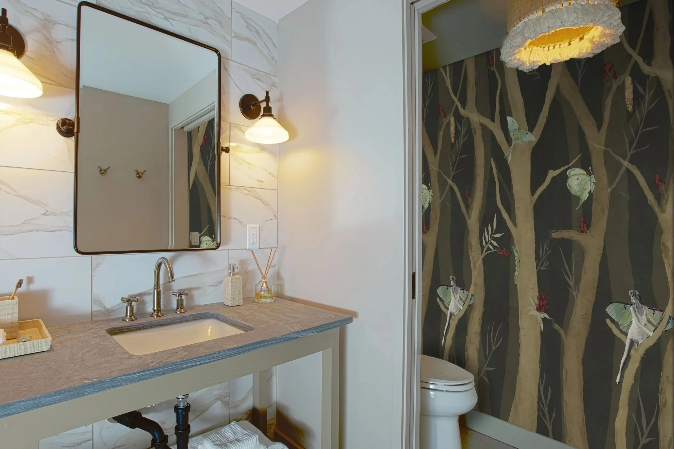 A bathroom with a marble countertop, a rectangular sink, and metallic faucets. A mirror is mounted on a wall with marble tiles. There are wall-mounted lights with shades above the mirror. The wall behind the toilet features dark wall paper with a tree and butterfly pattern. The bathroom also has a dispenser on the counter and a basket with a toothbrush. The flooring is light-colored, and there is a small white rug underneath the sink.