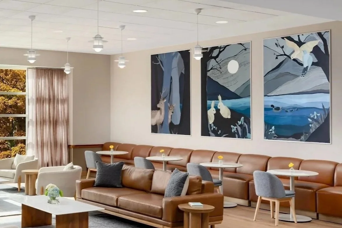A cozy lounge area with a large window showing autumn leaves outside, featuring beige curtains and a variety of seating including a light-colored armchair, white and gray chairs, and a long brown leather sofa. Small round tables with flowers are arranged throughout, and artwork depicting a woodland scene with deer and rabbits hangs on the wall.