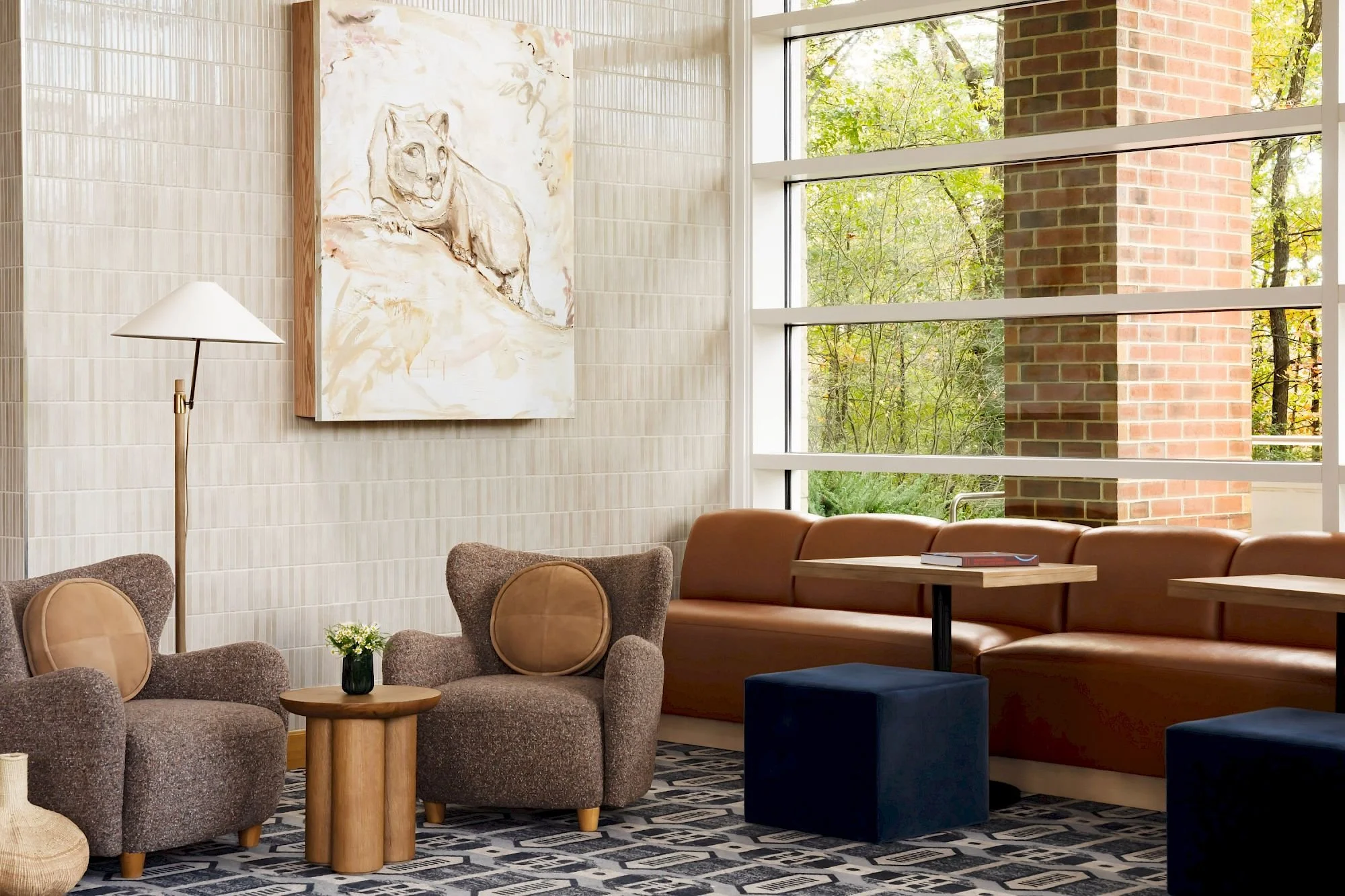 A cozy indoor sitting area with beige armchairs, a small wooden coffee table with a small plant, and a large tan sofa. There is a white wall with a textured pattern, a large abstract painting of a lion, a floor lamp with a white shade, and large windows revealing green trees and a brick wall outside.