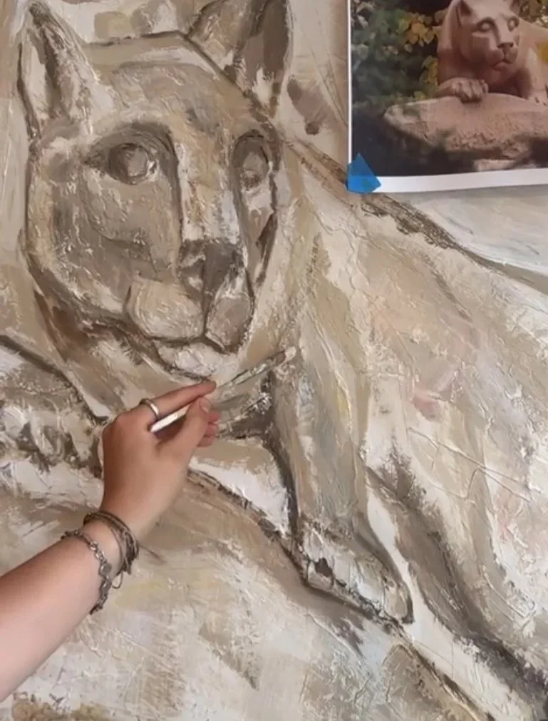 Artist painting a beige and brown animal portrait, possibly a big cat, on a textured wall, with a photo of a lion cub taped above.