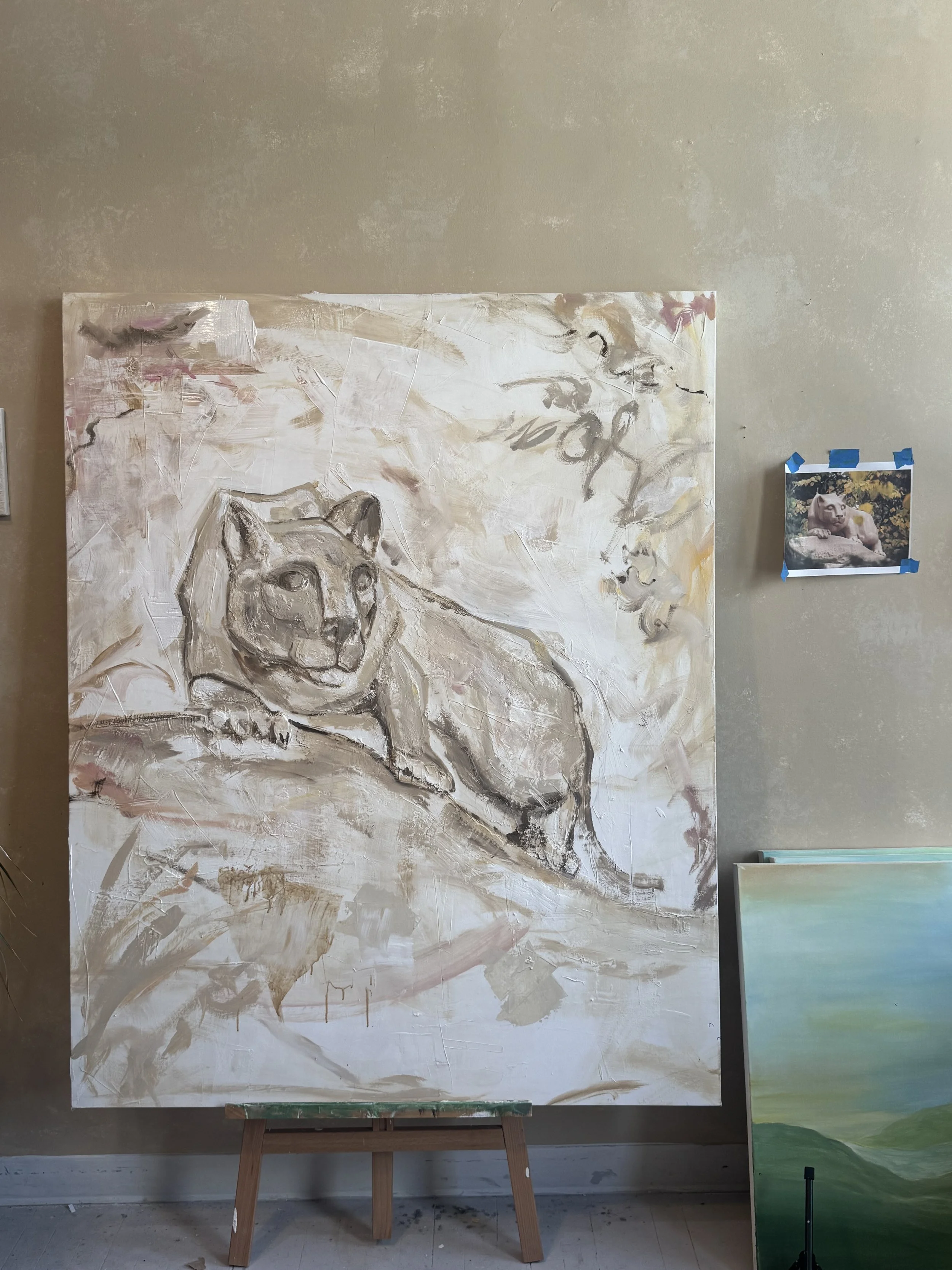 A large abstract painting of a lion's face in neutral and earthy tones, with expressive brushstrokes, propped on a wooden easel in an art studio, with a smaller landscape painting leaning against the wall.