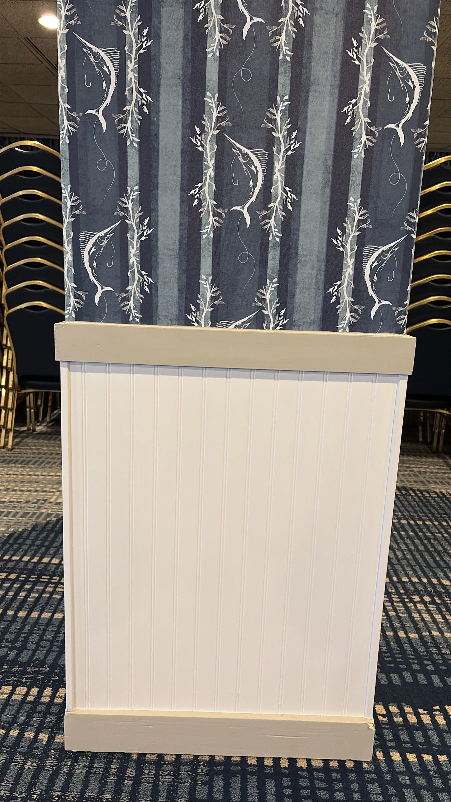 White panel with vertical grooves and a trimmed top and bottom, placed in front of a blue wall with a tree and fish pattern, beside stacked black chairs with gold trim in a conference or event space.