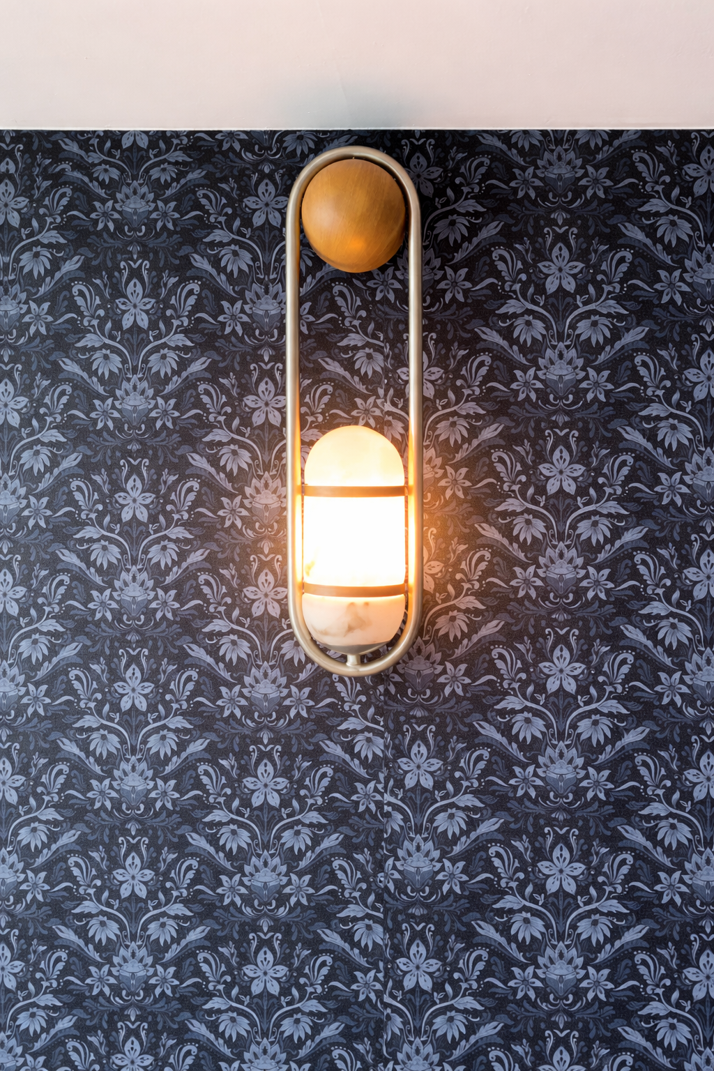 Wall-mounted modern lamp with a round wooden top and a glowing white spherical bottom, against a dark floral-patterned wallpaper.