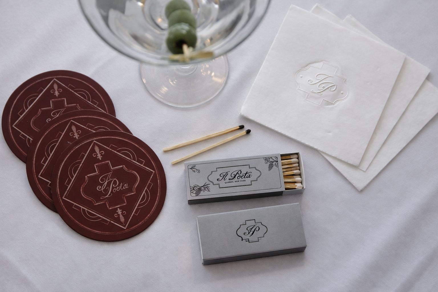 A table setting with a glass containing green olives on a skewer, three burgundy coasters with an intricate design and the words 'A Poeta,' two white cloth napkins with a monogram, two boxes of matches labeled 'A Poeta,' and a black and white box of matches on a white tablecloth.