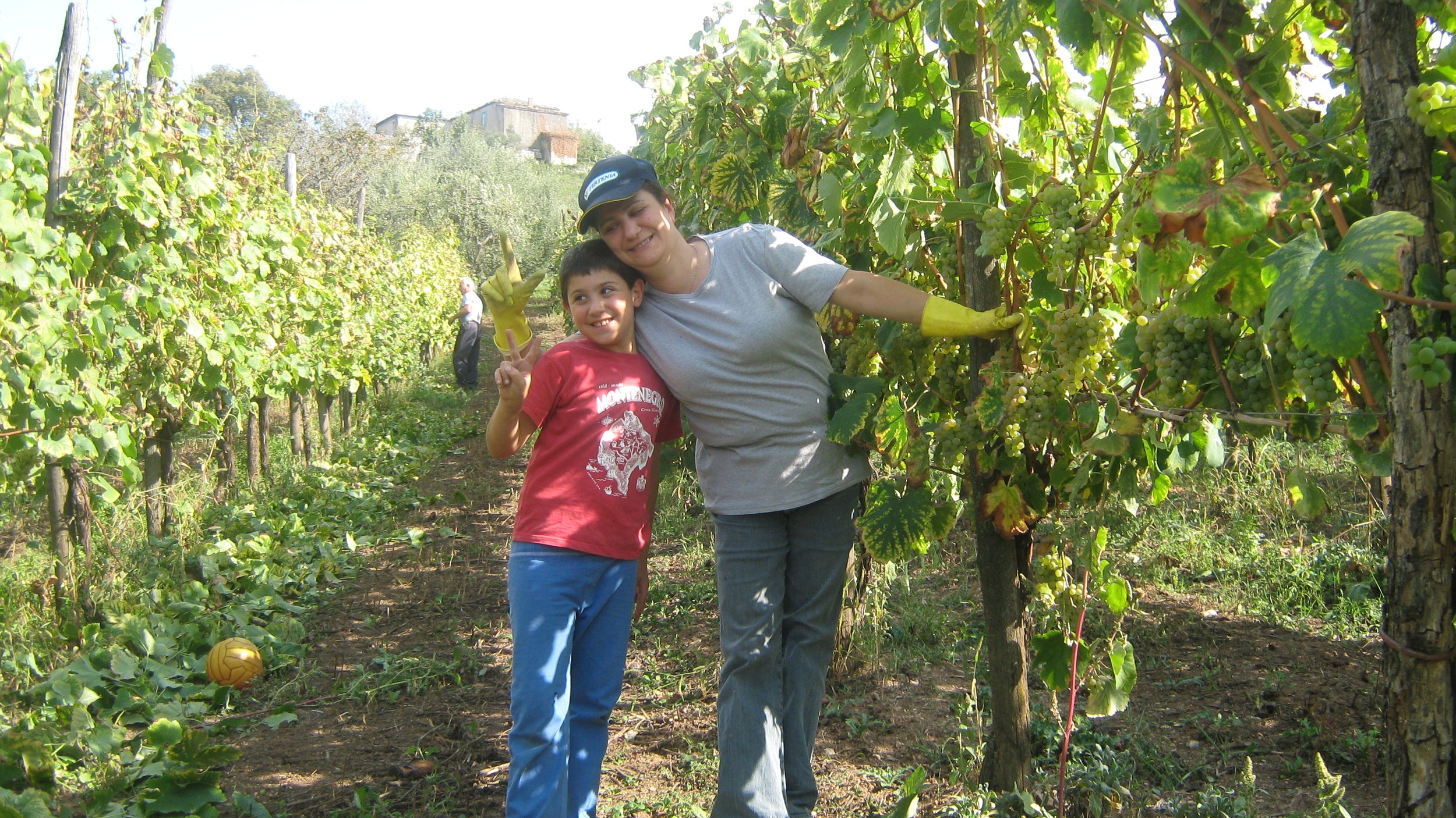Un bambino e una donna sorridenti in un vigneto, che fanno il segno della pace e raccolgono uva.