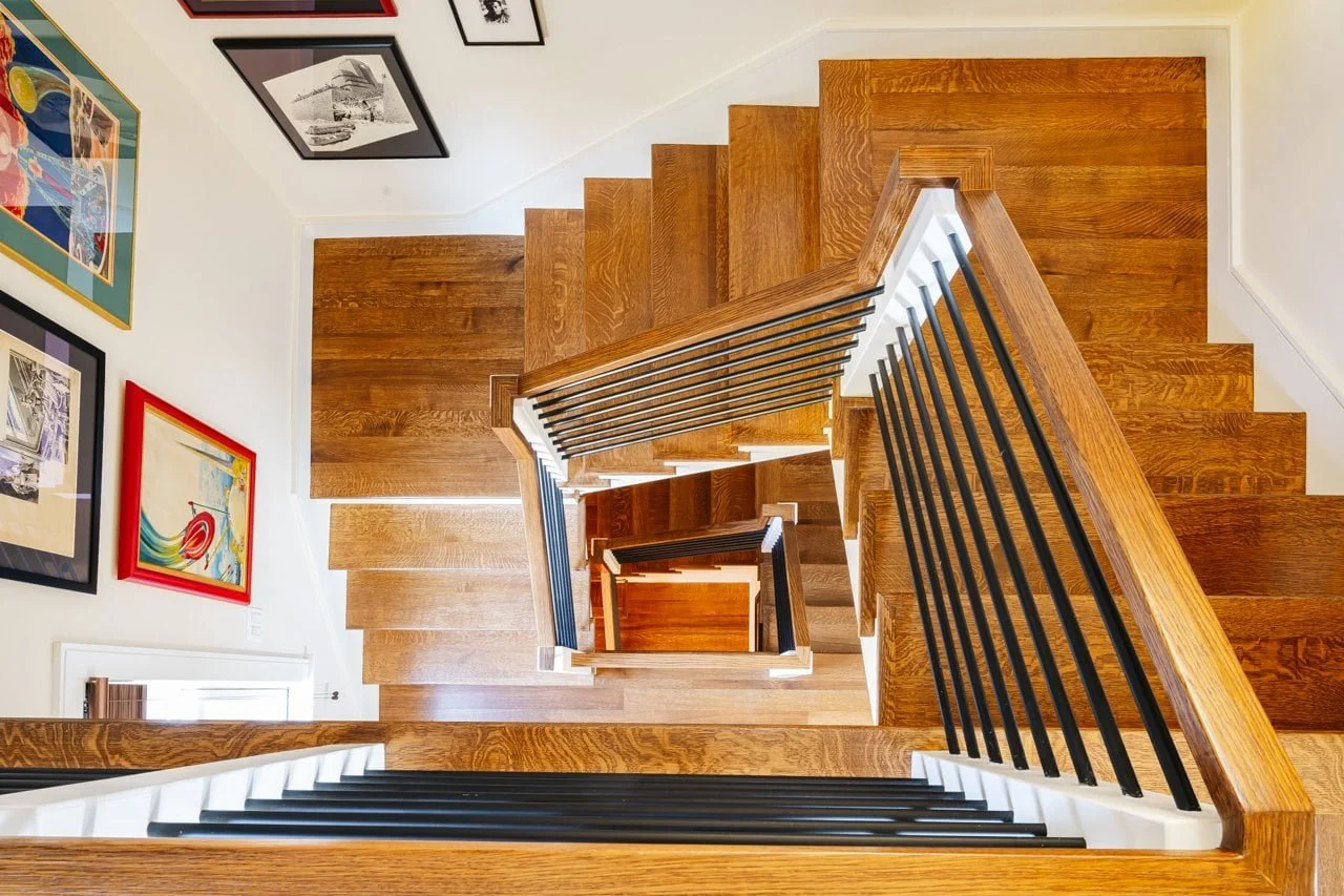 View from the top of a spiral staircase looking down to the lower floors, with wooden steps and black metal railings, decorated with framed art on white walls.