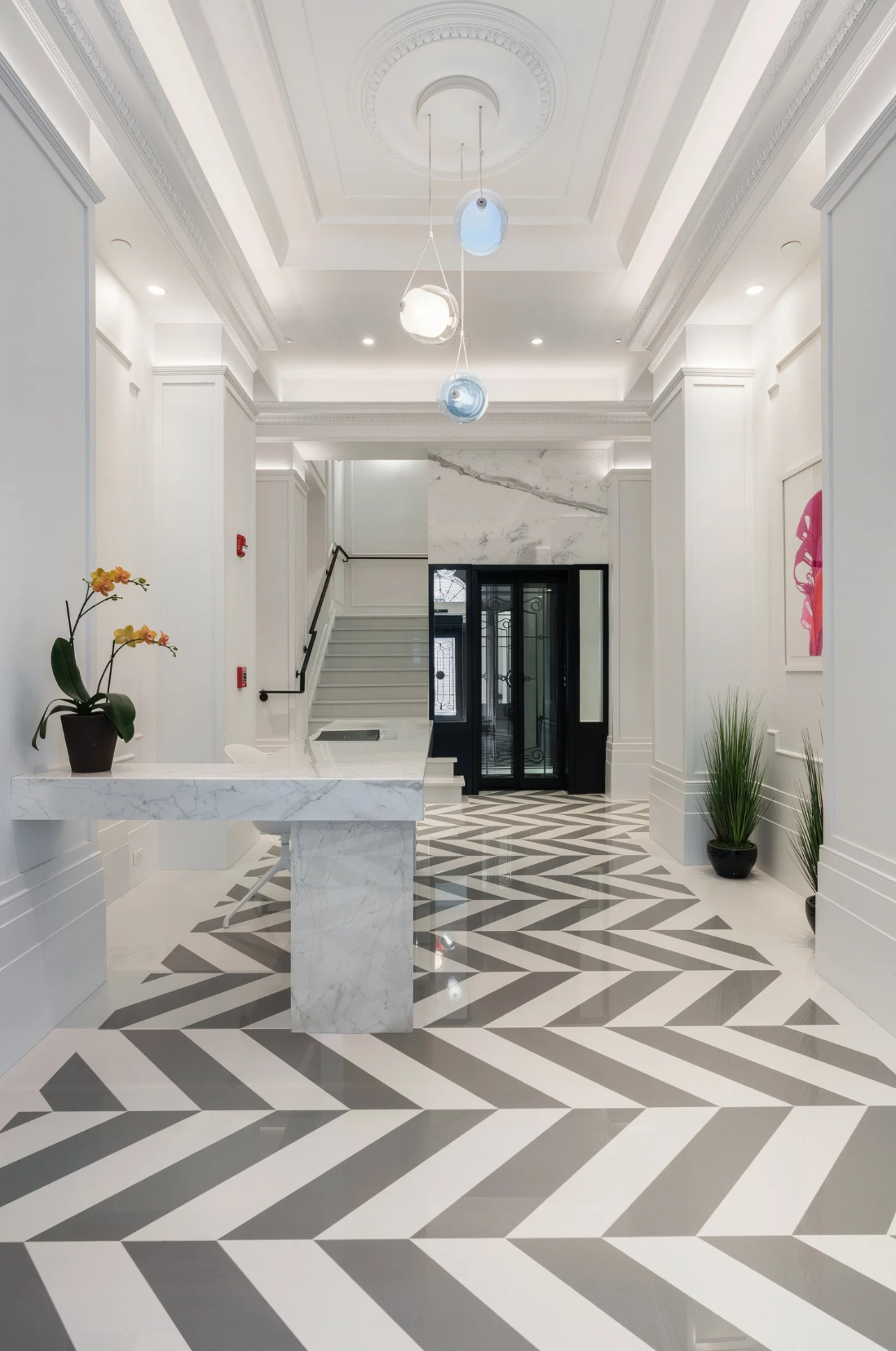 Modern hotel lobby with white walls, a patterned black and white floor, a marble reception desk, potted plants, an art piece, and a staircase leading upstairs.