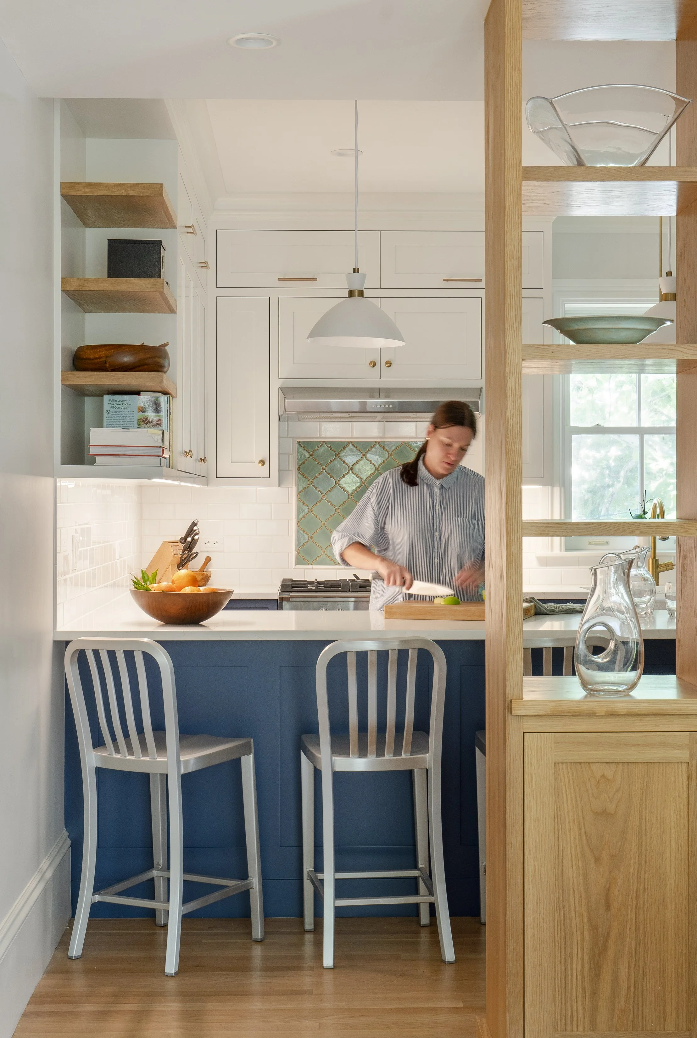 A person preparing food at a kitchen counter with a bowl of oranges on the table, white cabinetry, and open shelving with decorative bowls, in a bright kitchen with a window and modern decor.