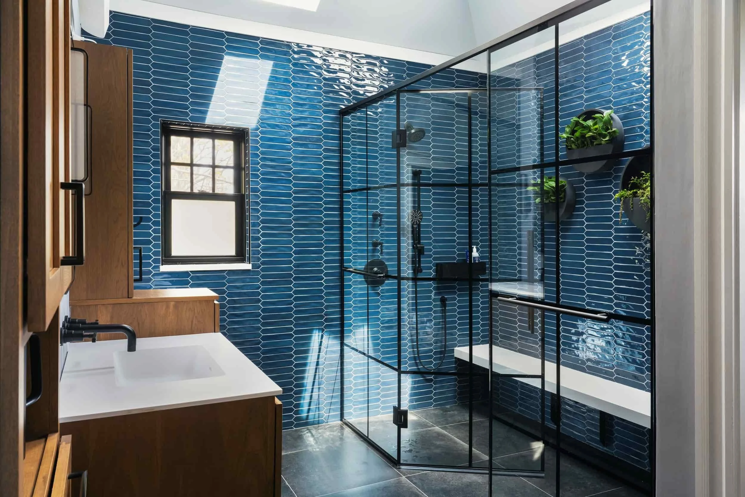Modern bathroom with a black-framed glass shower enclosure, blue hexagon tile walls, a white vanity with a black faucet, black floor tiles, and a small window letting in natural light, decorated with green plants.