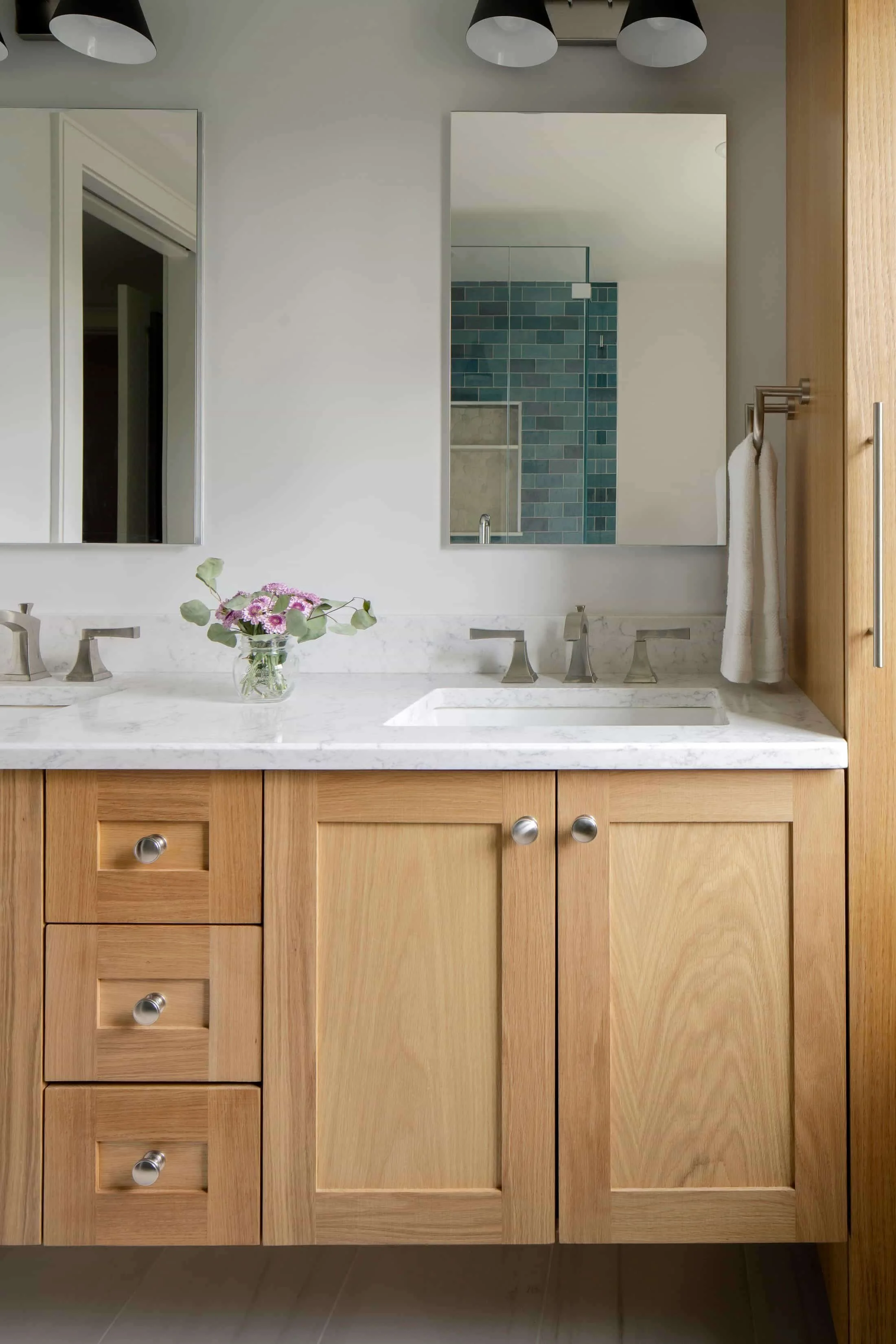 Wooden bathroom cabinet with silver knobs, white marble countertop, and two undermount sinks, with a small vase of pink and purple flowers.