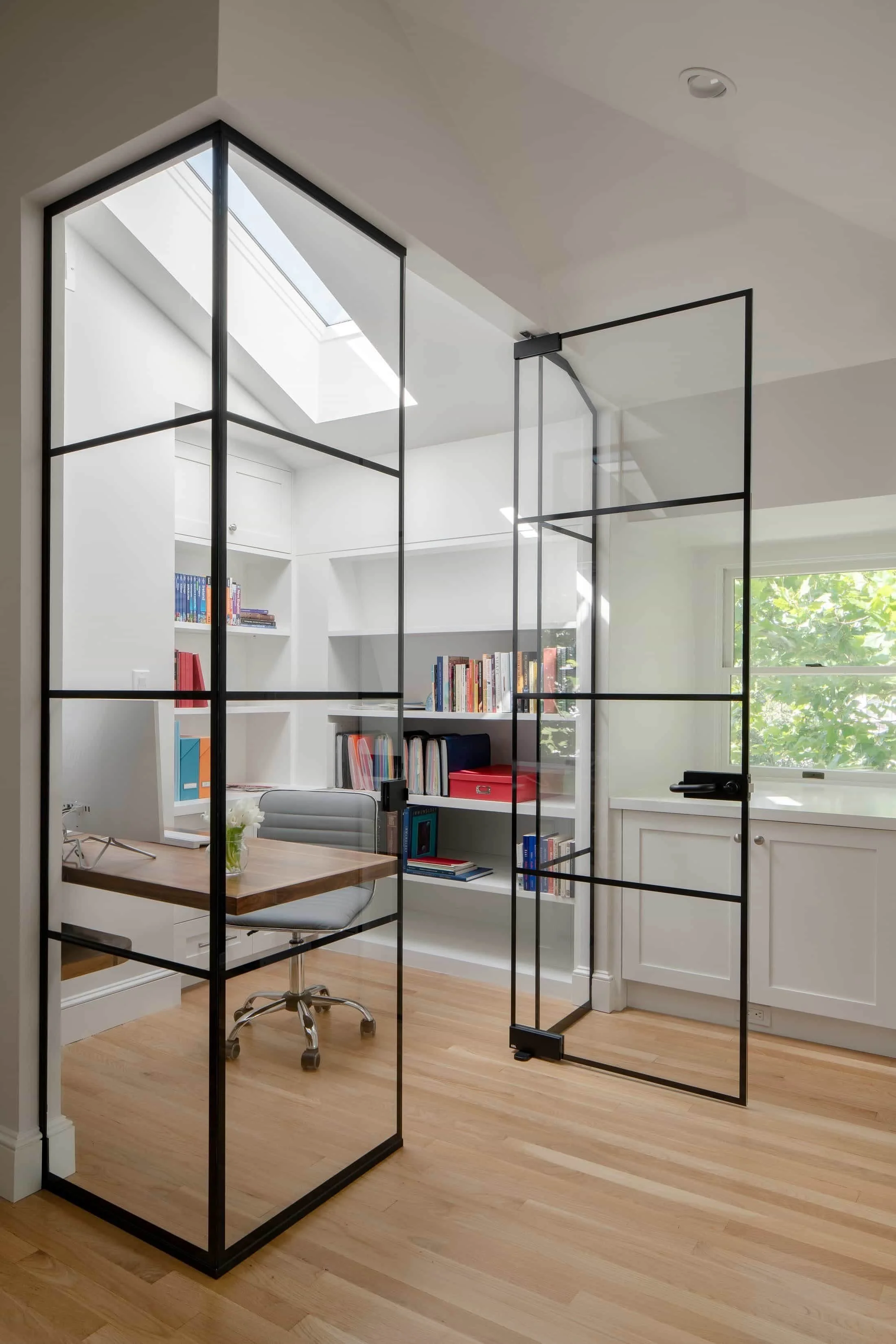 Modern home office with a glass and metal room divider, a wooden desk, a gray swivel chair, bookshelves on white walls, a window, and hardwood floors.