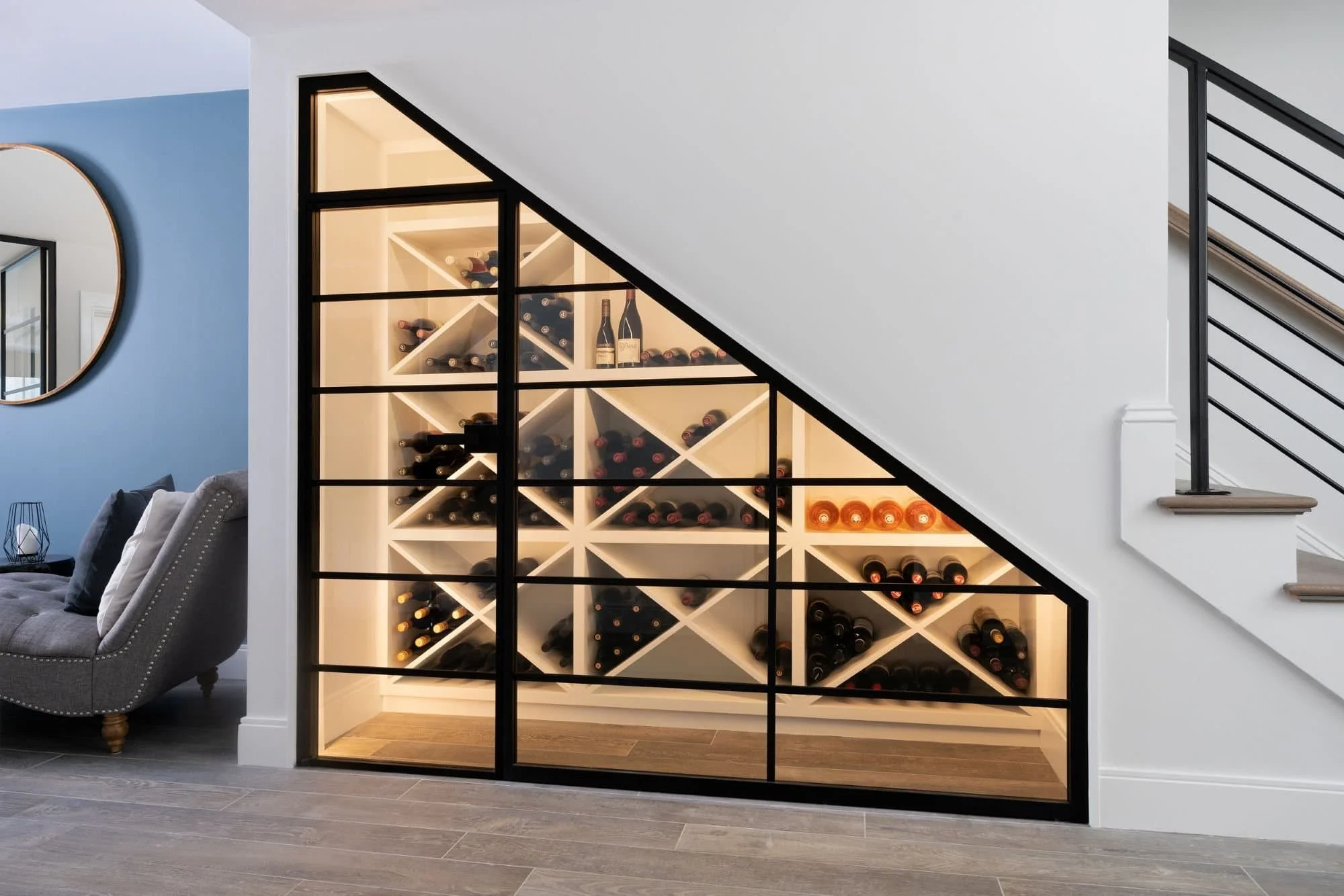 A modern wine cellar under stairs with wood and black metal framing, filled with wine bottles stored horizontally and illuminated with warm lighting, in a contemporary living space.