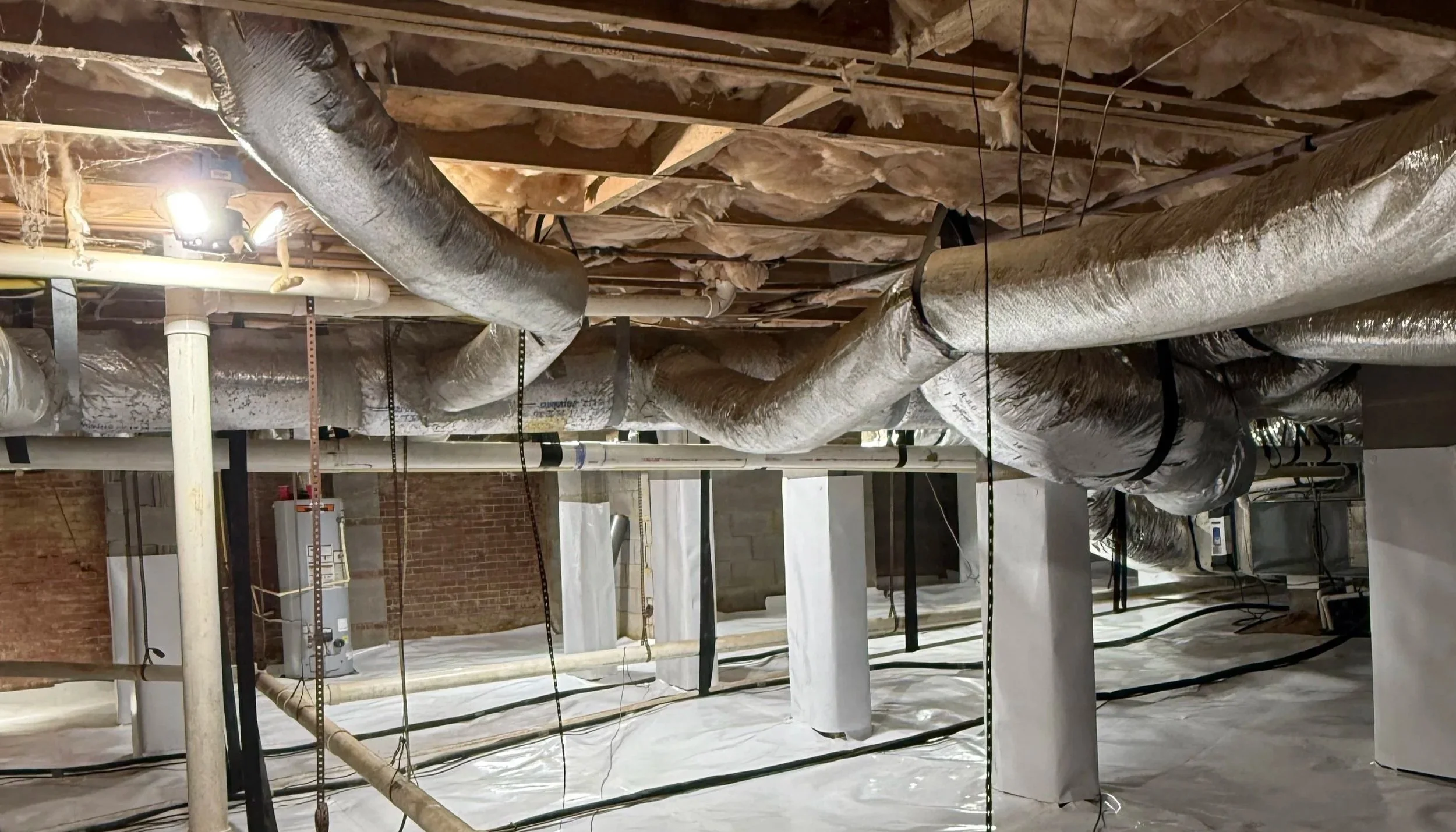 Construction site ceiling with exposed pipes, ducts, and insulation. Various pipes run across the ceiling, some wrapped in insulation. The ceiling joists are visible, and the space is partially finished with bricked walls and construction materials.