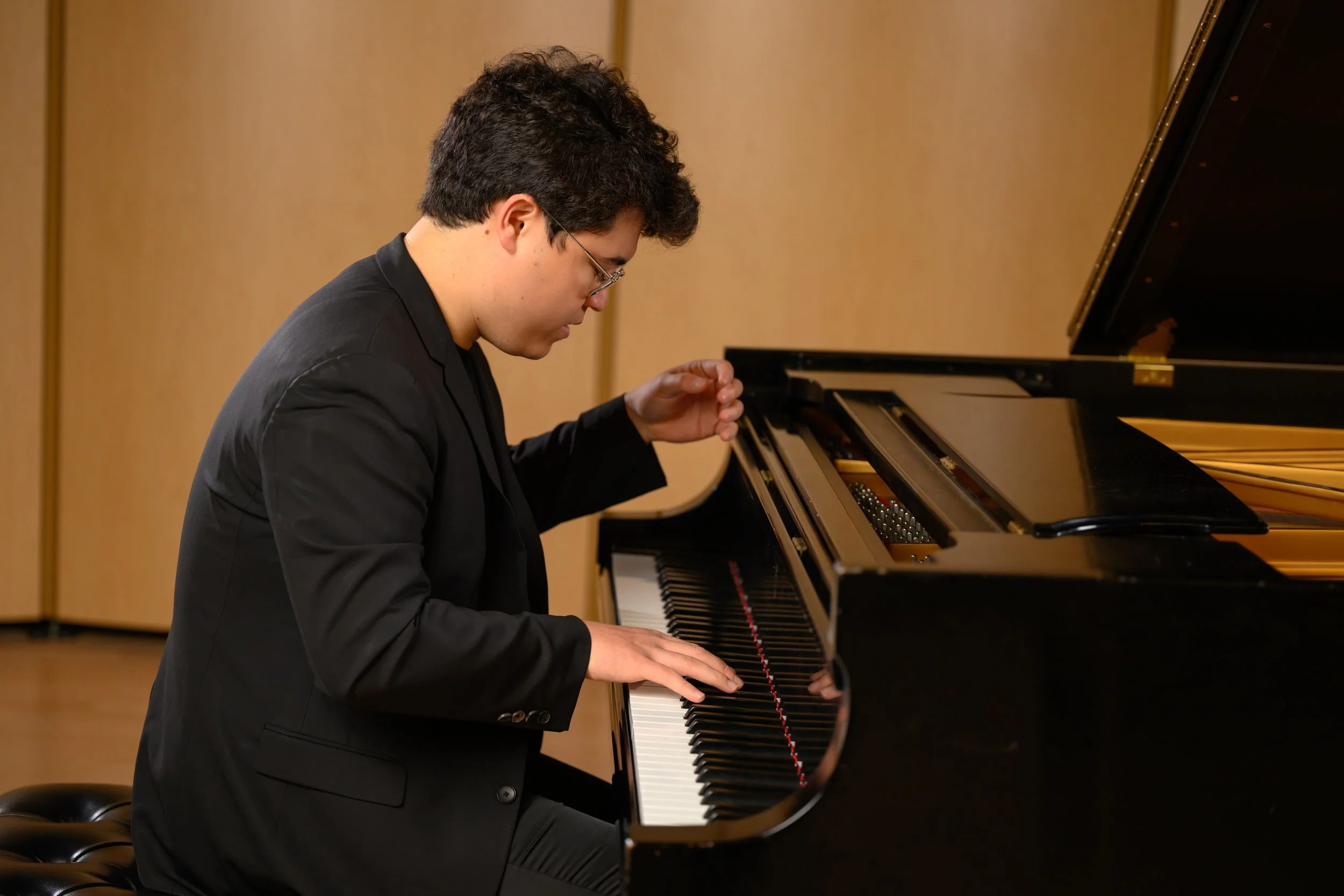 A young man in a black suit playing a grand piano in a room with wood paneling.