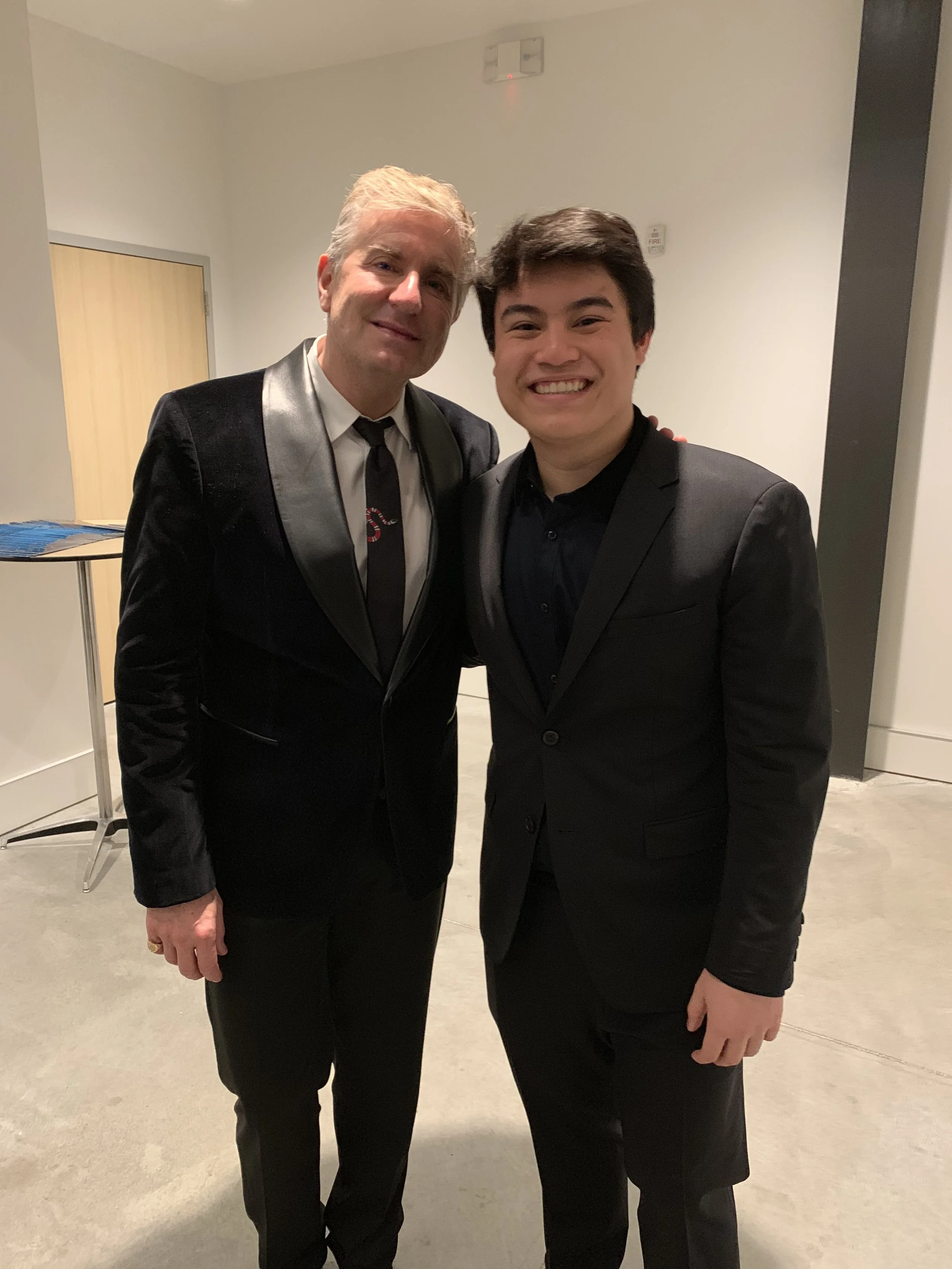 Two men dressed in formal black suits smiling and posing together indoors.