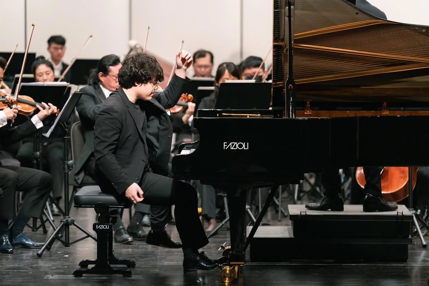 A young pianist in a black suit performing on a Fazioli grand piano with an orchestra in the background.