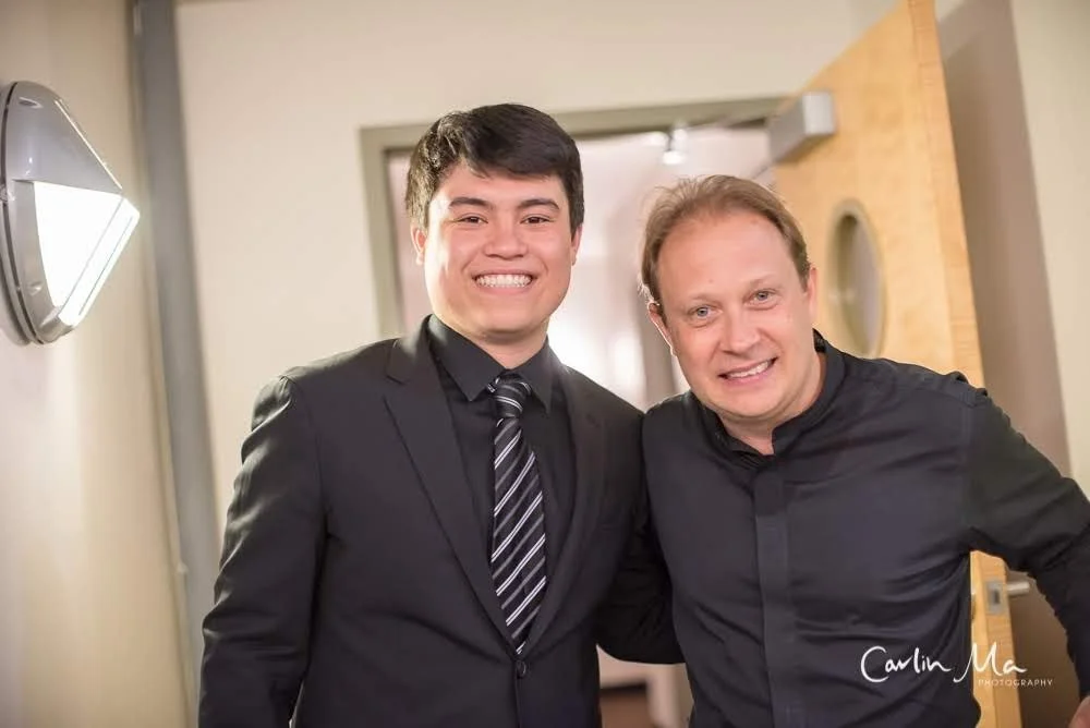Two men smiling and standing close together indoors, one in a black suit with a striped tie, and the other in a black shirt, in front of a beige wall with a wooden door.