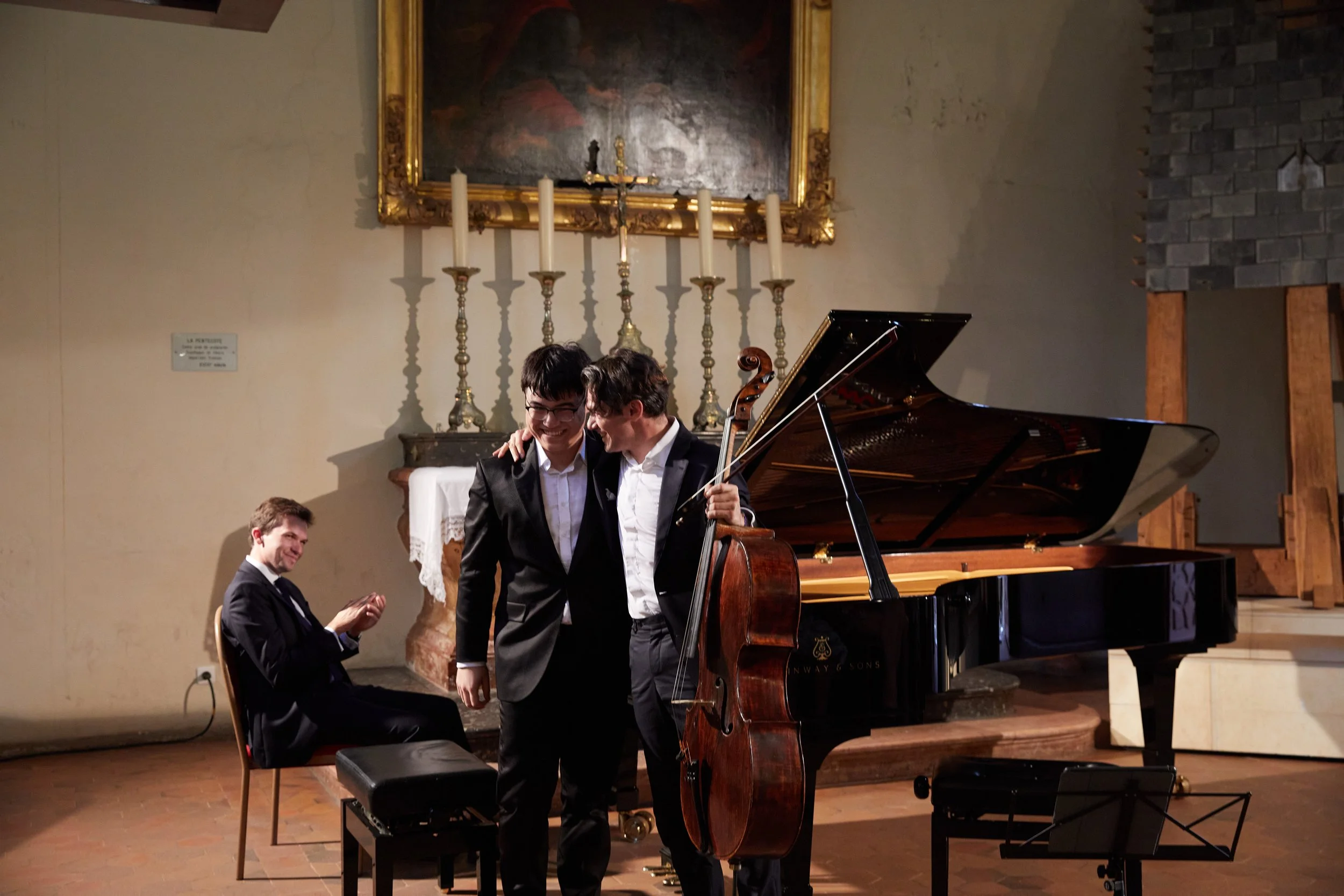 Three musicians, two standing and one sitting, in a room with a grand piano, candles, a large painting, and a brick fireplace. The two standing musicians are smiling and embracing, one with a violin and the other with a piano.