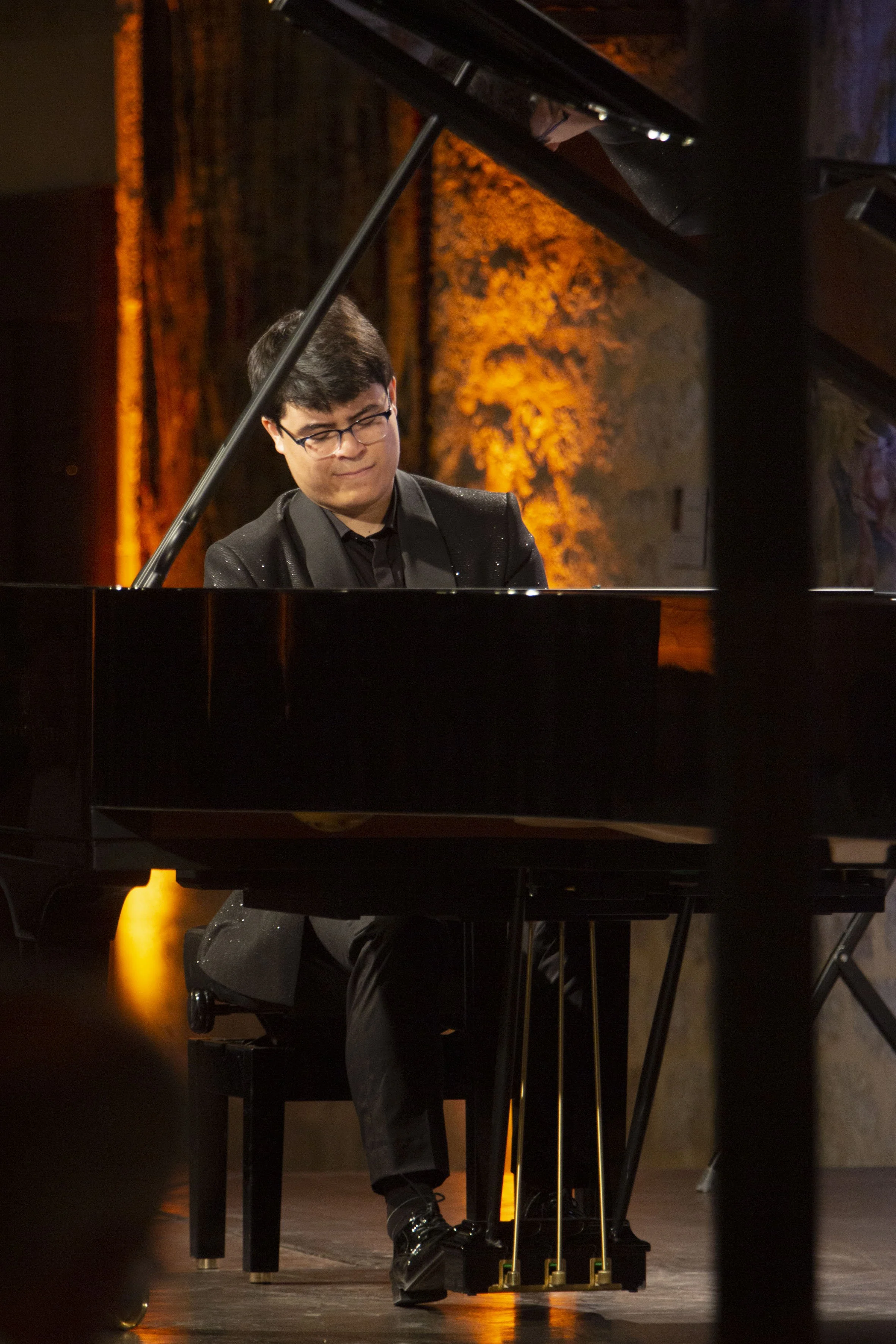 A man playing a grand piano on stage, dressed in a black suit and glasses, with a warm, orange-lit background.