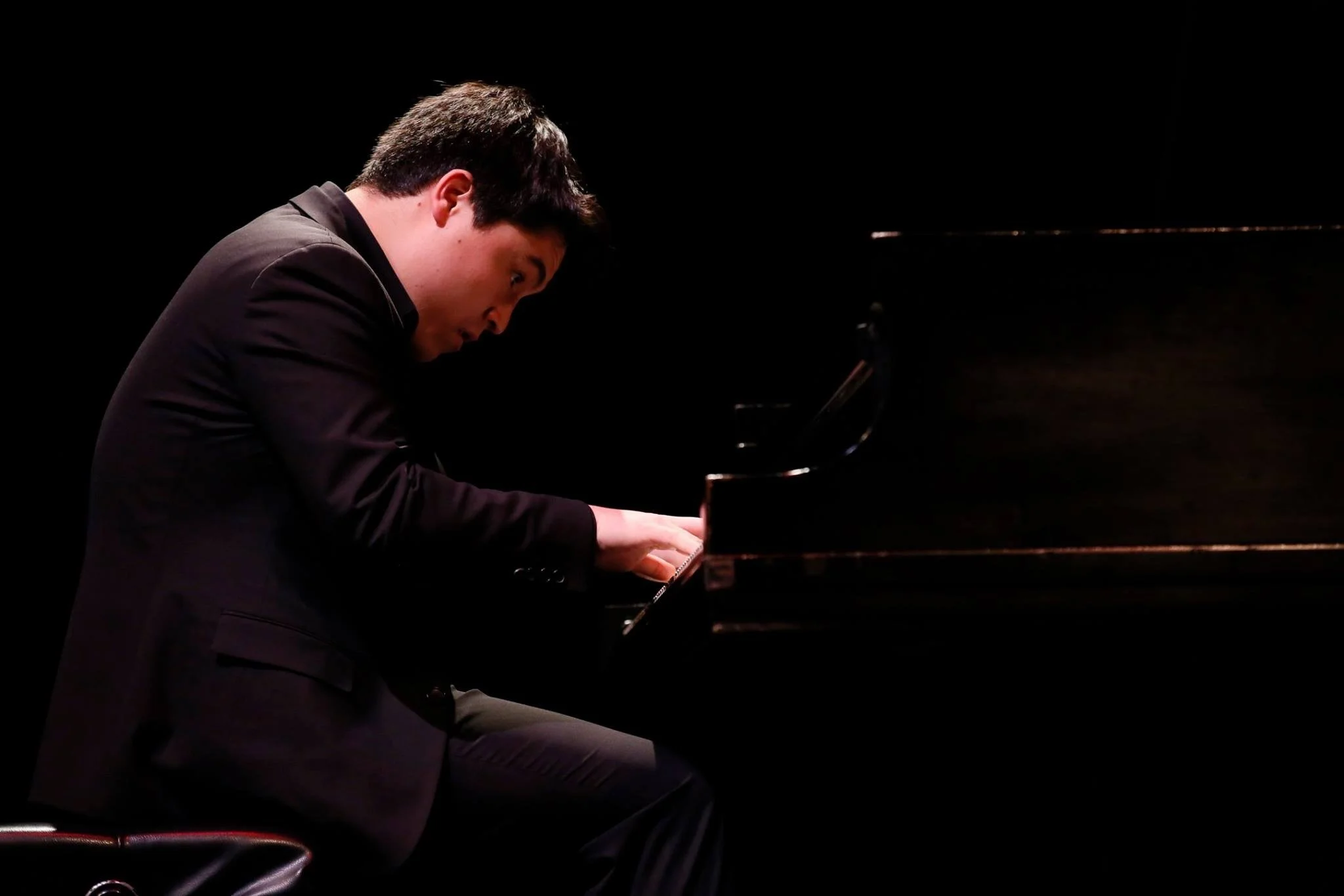 A man in a black suit playing a grand piano on a dark stage.