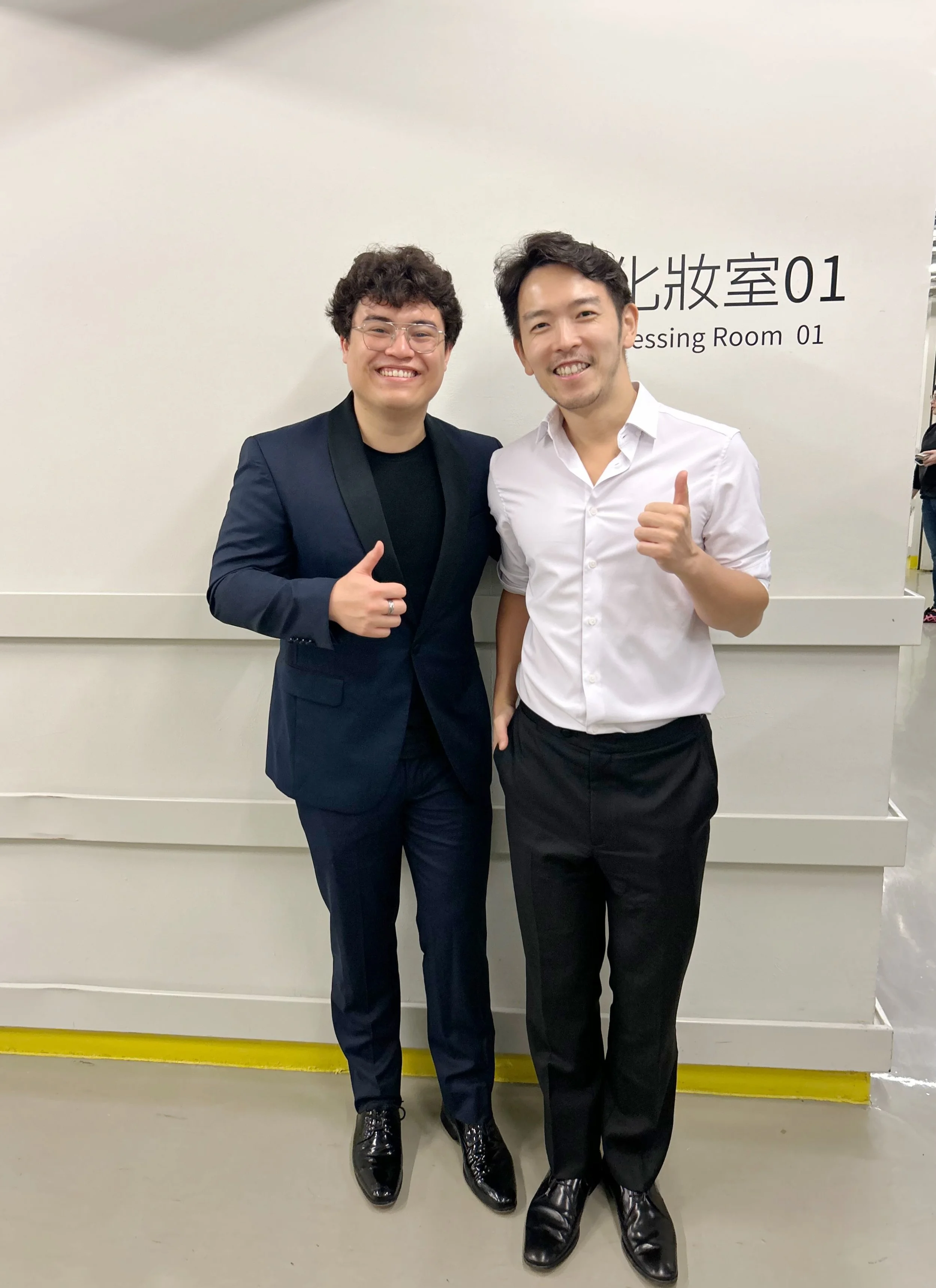 Two men smiling and giving thumbs up, standing in front of a sign that says 'Dressing Room 01' in English and Chinese.