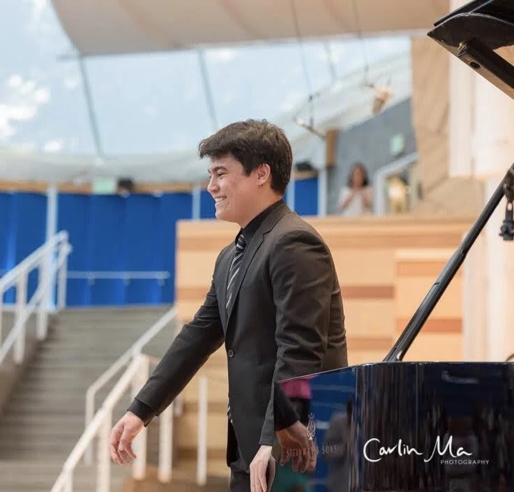 A young man in a black suit smiling near a dark blue grand piano inside a modern building with wooden accents and stairs.
