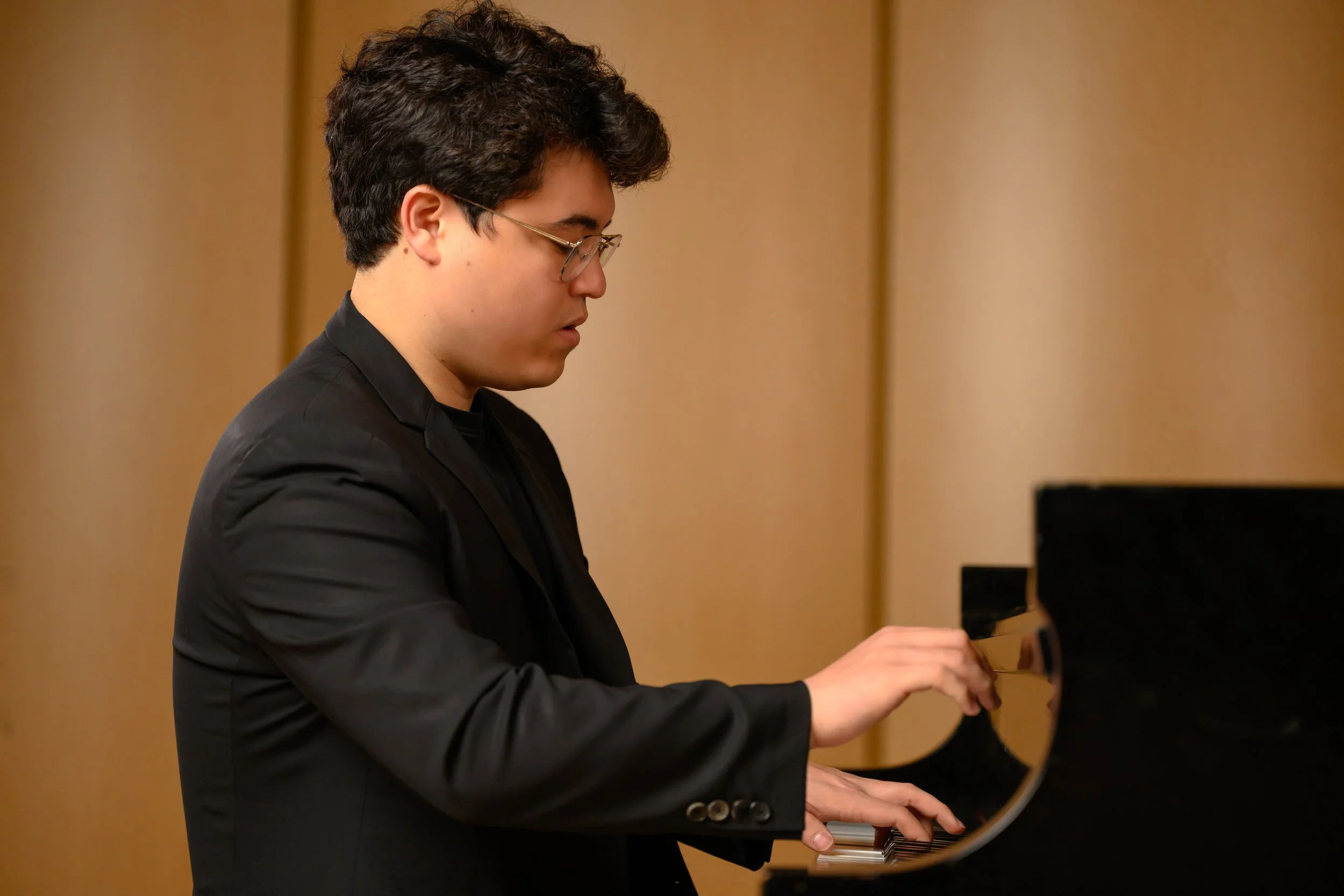 A young man with glasses and dark hair playing a grand piano.