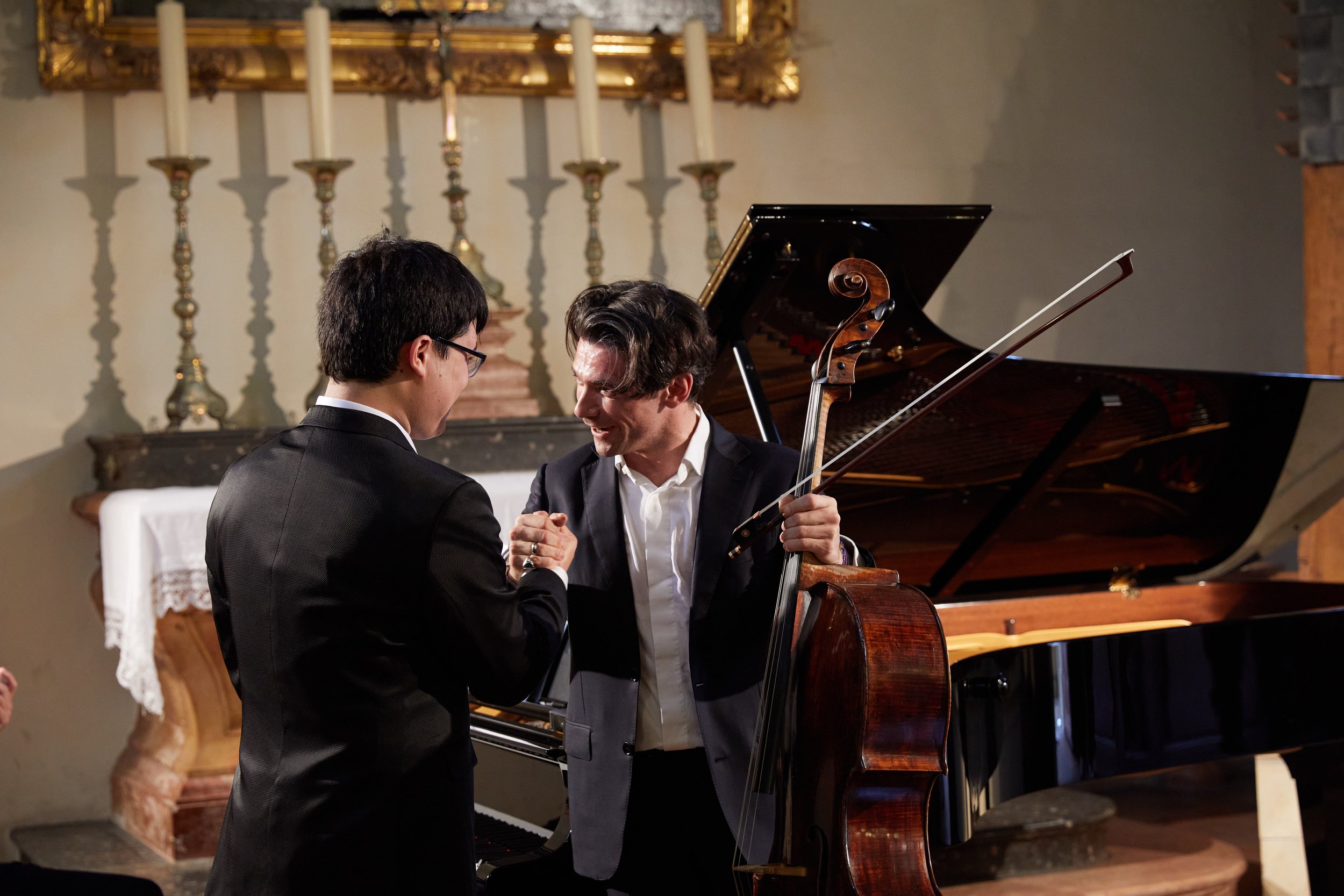 Two men in formal attire shaking hands or sharing a moment beside a grand piano and a cello in an elegant room with candlesticks and a framed mirror.