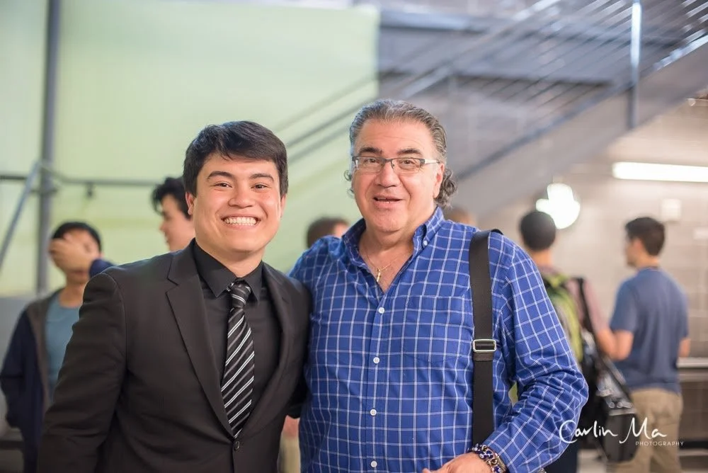 Two men, one young in a black suit and tie, and one older in a blue checkered shirt, smiling together in a public space with other people in the background.