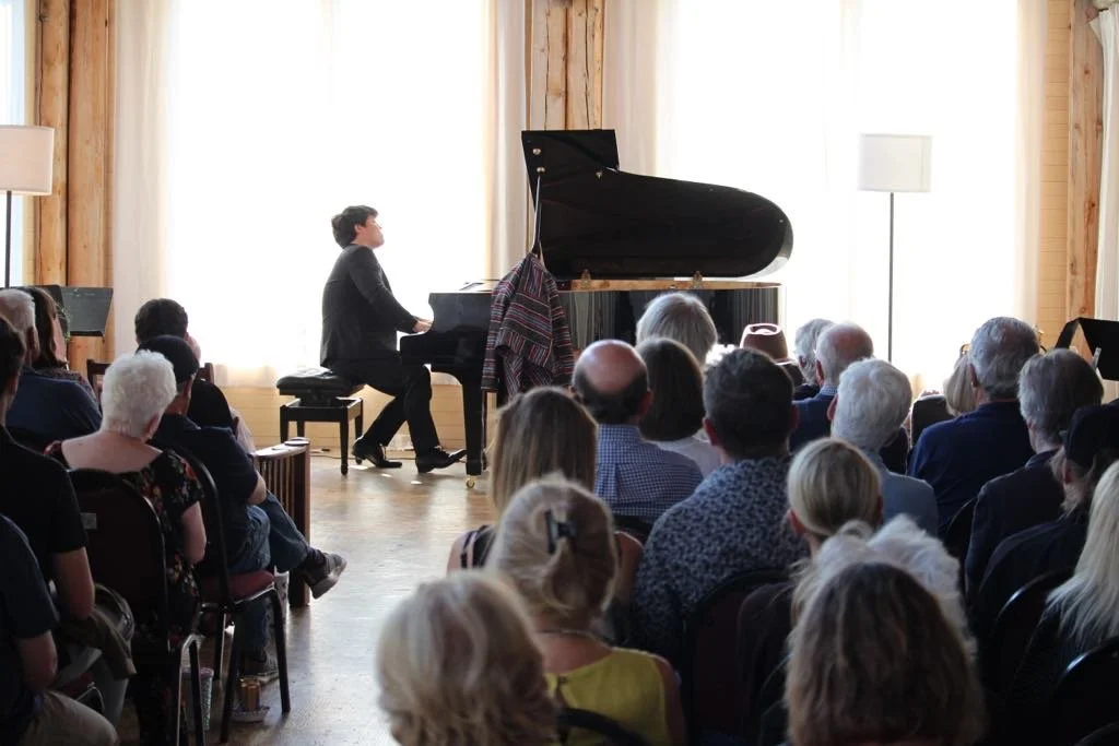 A man playing the piano in front of an audience in a room with wooden walls and large windows letting in natural light.