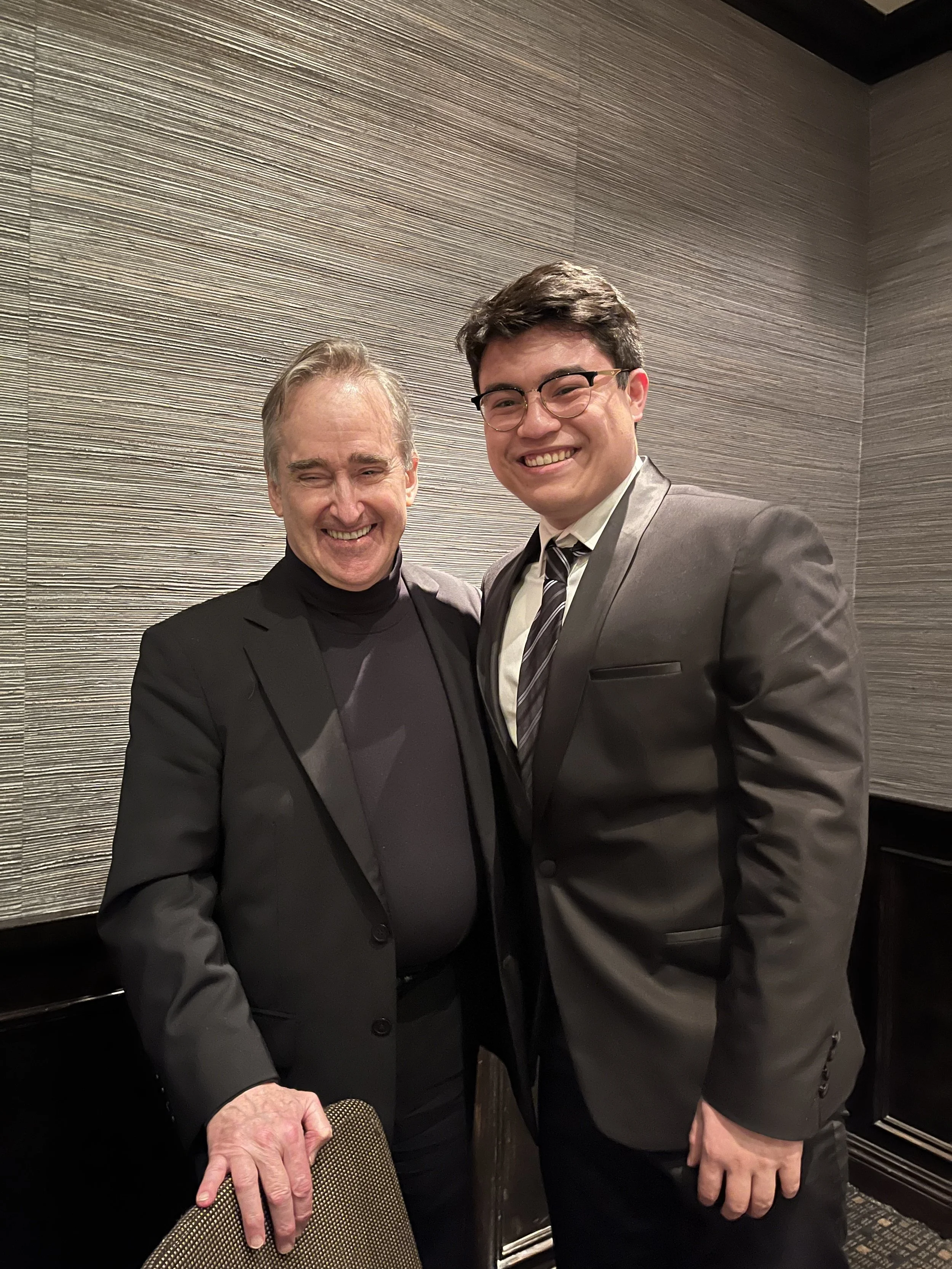 Two men dressed in suits smiling and standing close together in front of a textured wall, one resting his hand on a chair.