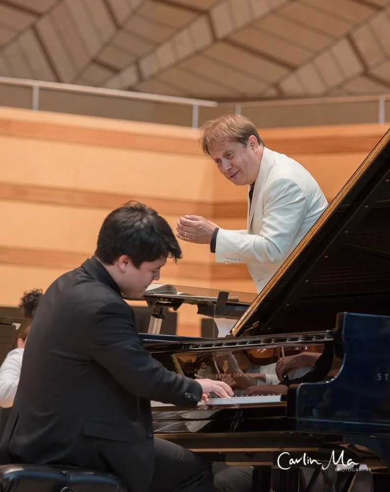 A young man playing the piano while an older man in a white suit wearing a black shirt conducts or instructs him inside a wood-paneled room or auditorium.