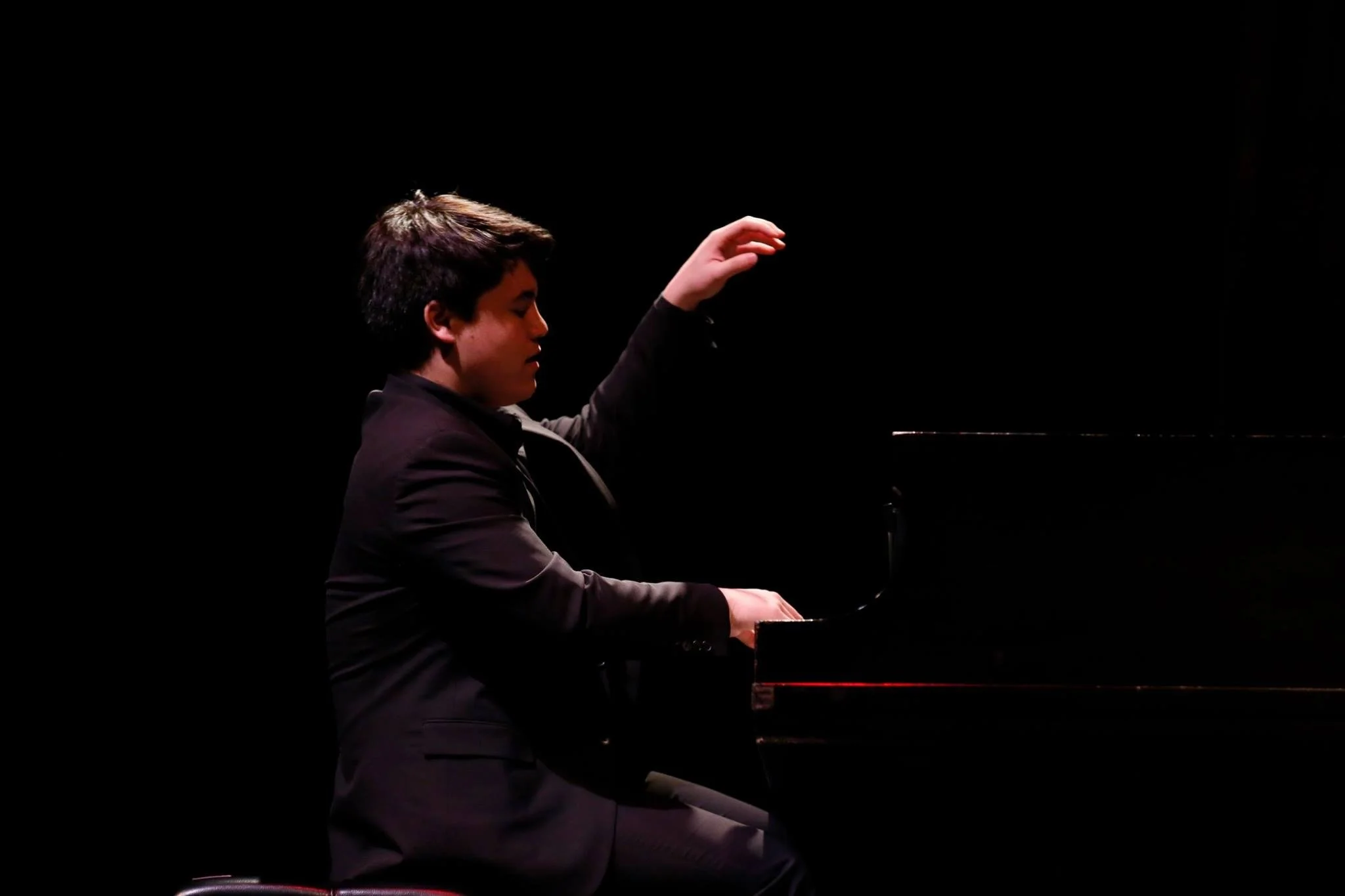 A young man in a suit playing a grand piano on a dark stage.