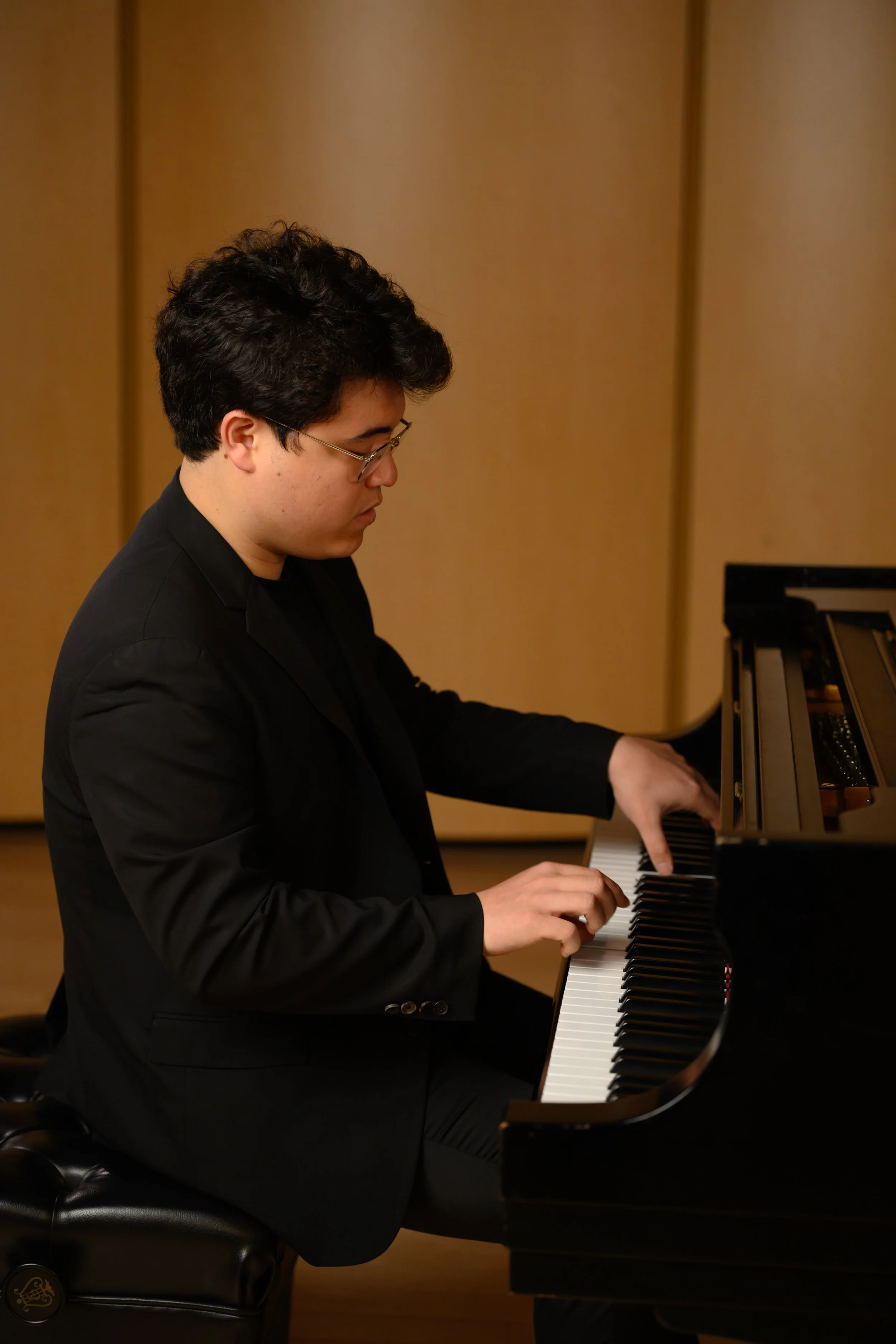 A young man with dark hair and glasses playing a black grand piano in a room with beige walls.