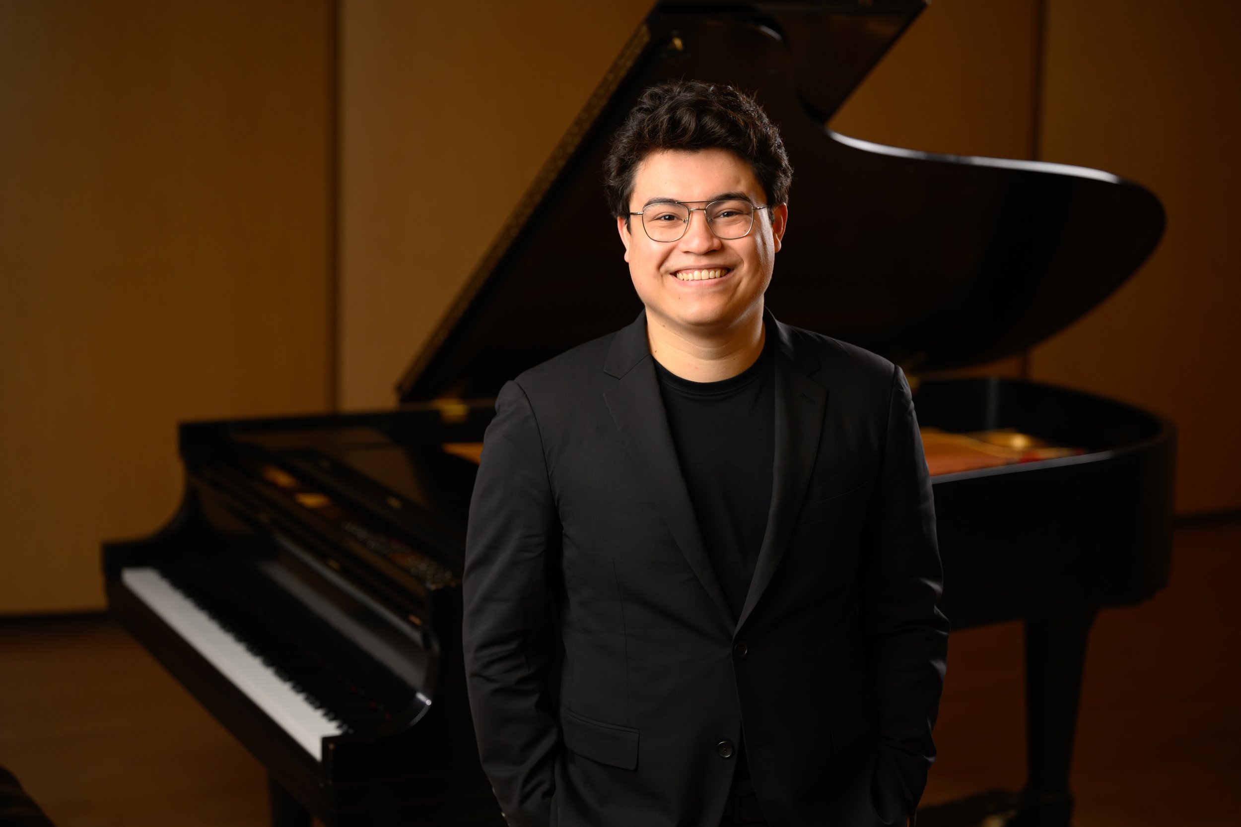 Young man with glasses in a black blazer standing in front of a grand piano in a music studio.