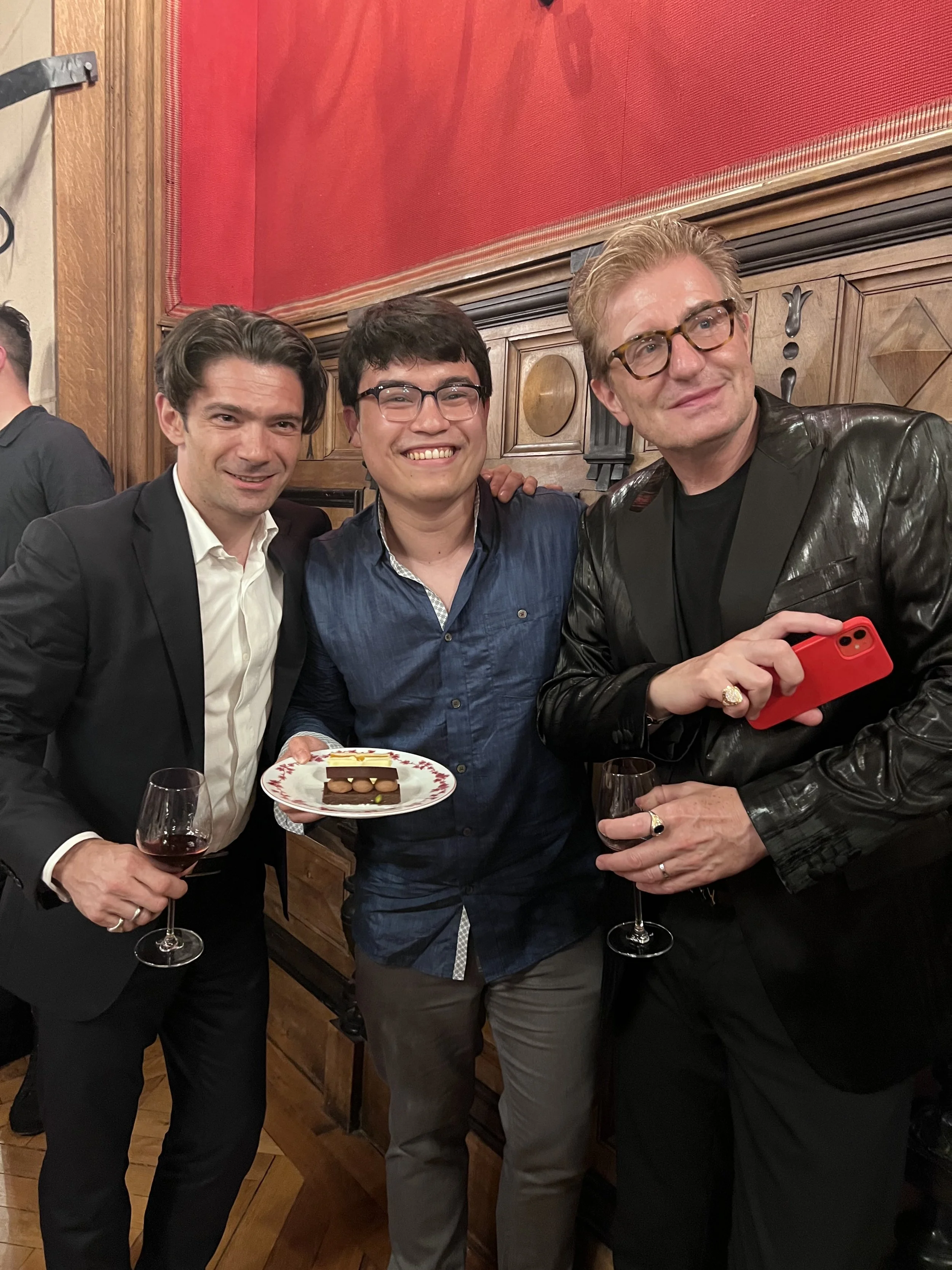 Three men at a celebration, holding glasses of wine, with one man holding a plate of dessert, in a room with wooden paneling and red walls.