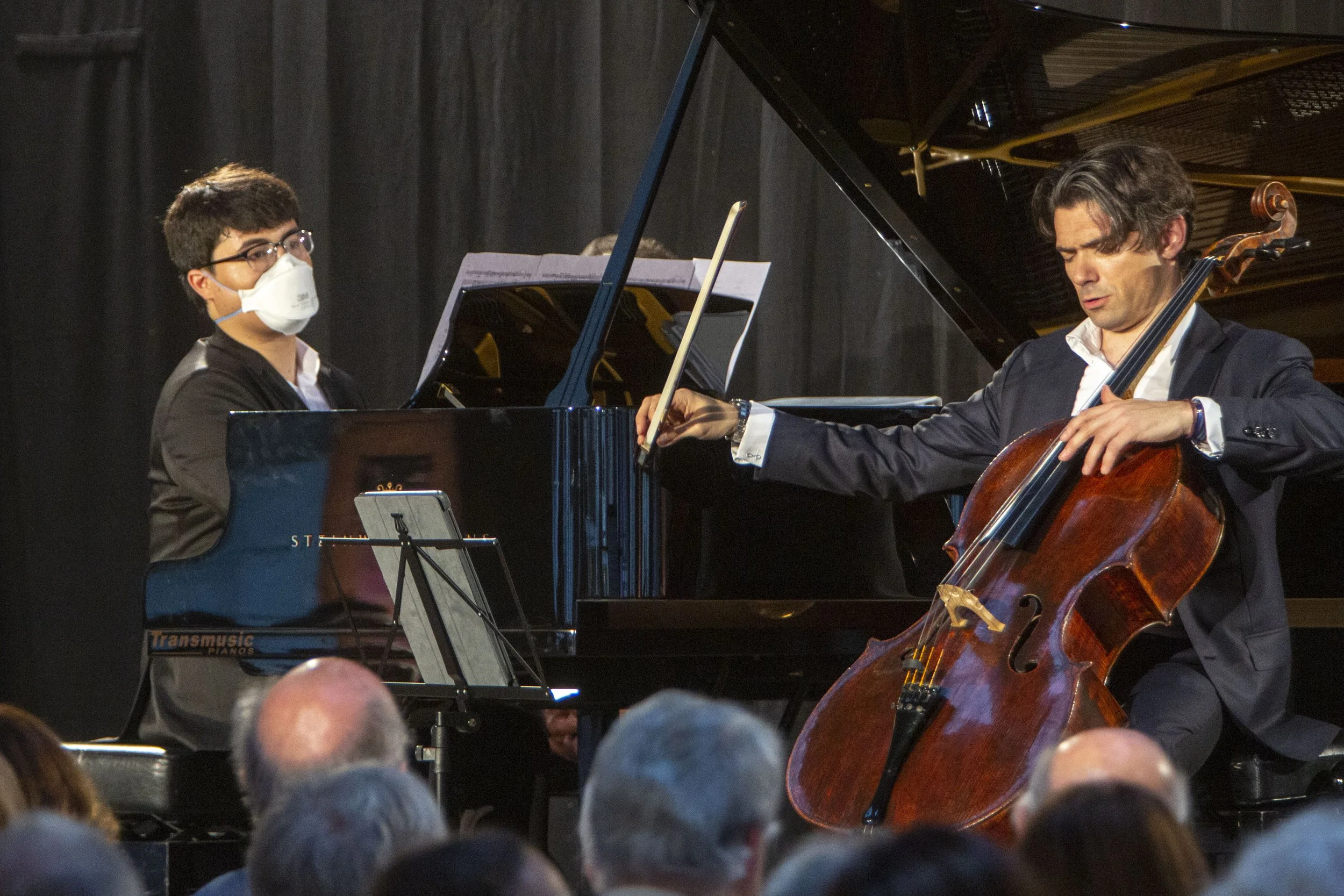 Two musicians performing on stage: a young man wearing glasses and a face mask playing the piano, and another man in a suit playing the cello. Audience visible in foreground.